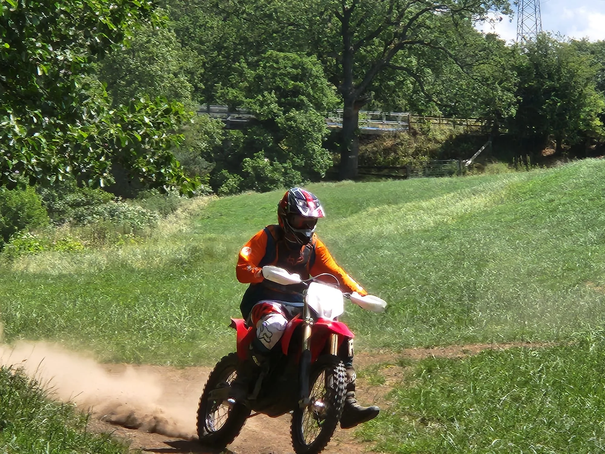 A person wearing an orange jacket and a black helmet riding a small red dirt bike on a dirt trail in a lush green park with trees and bushes.