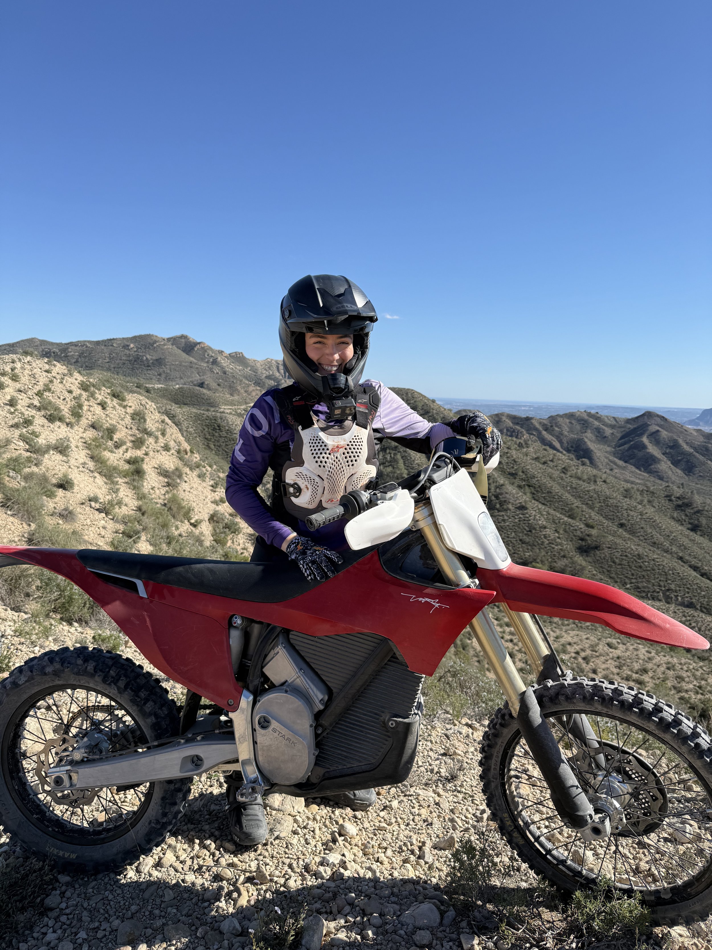 A woman in motocross gear, including a helmet and riding suit, sitting on a red and white electric dirt bike on a rocky trail in a mountainous desert landscape with a clear blue sky.