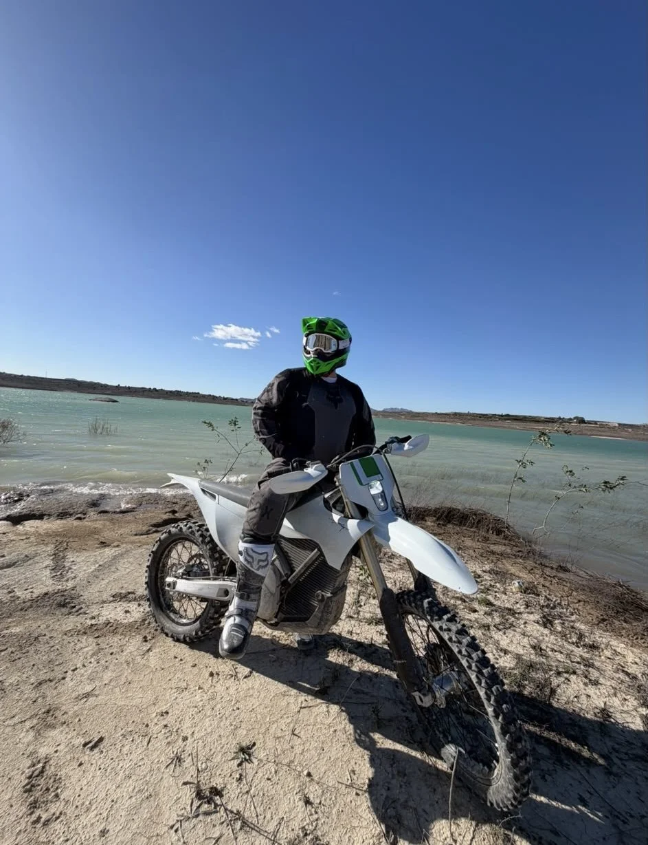 Person wearing a green helmet and black riding gear sitting on a white dirt bike by a lake with clear blue sky.