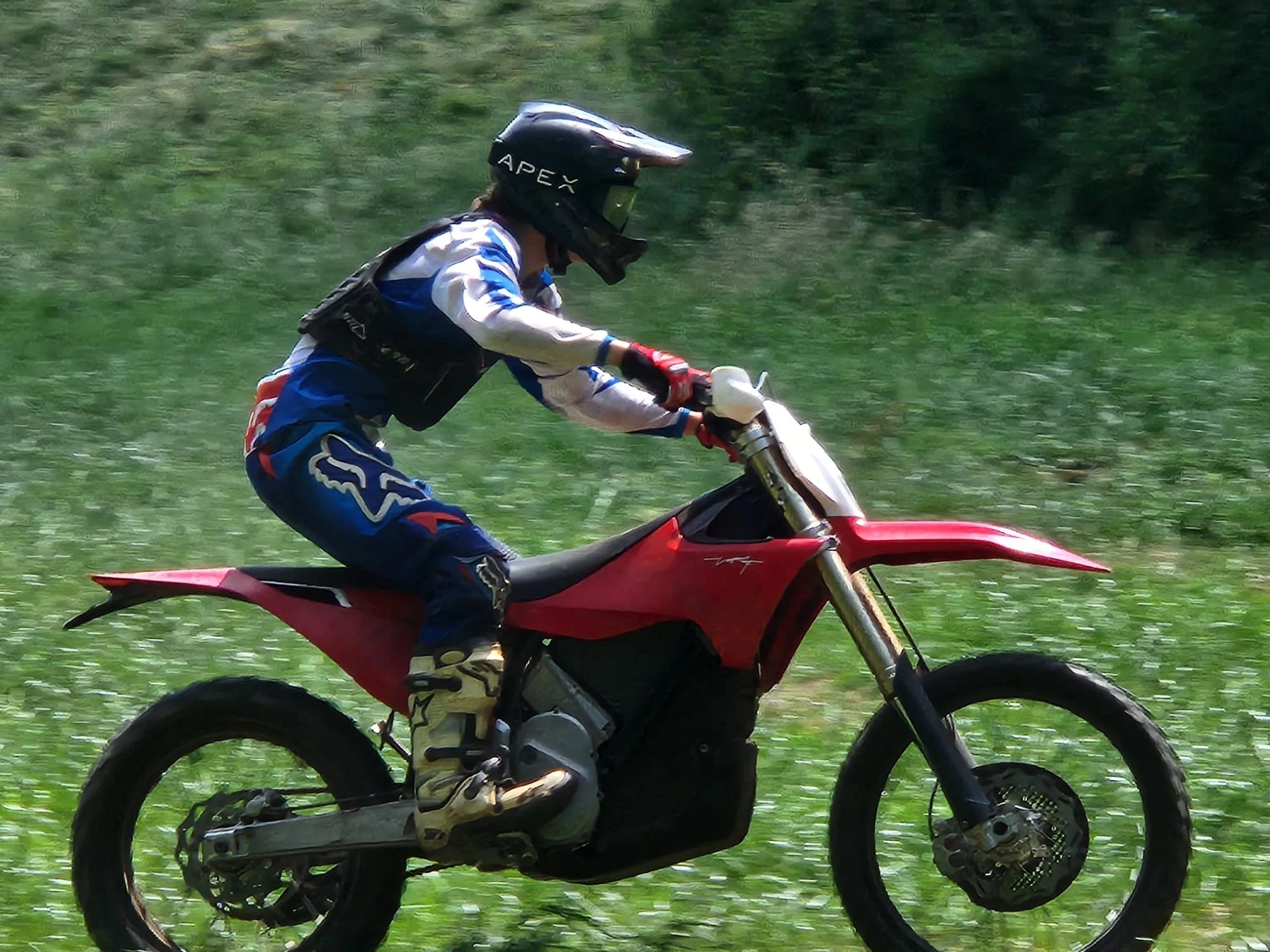 A person riding a red dirt bike off-road on green grass while wearing a full face helmet and motocross gear.