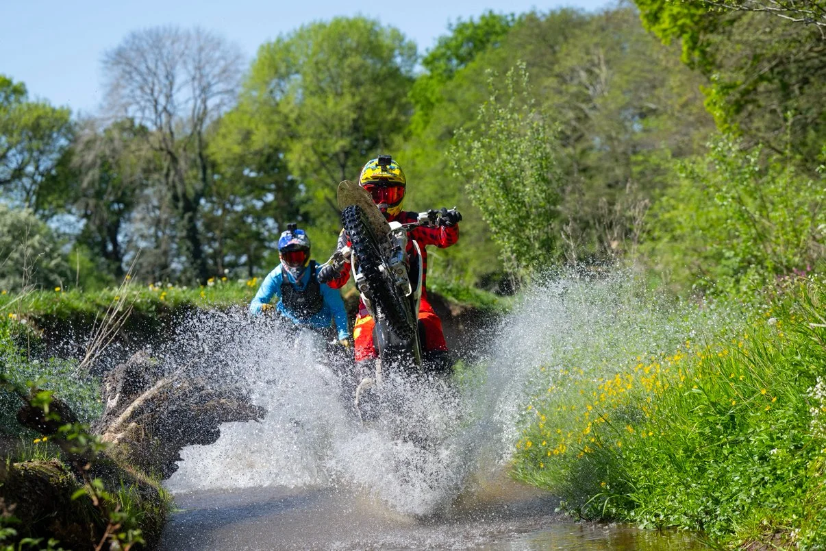 Two dirt bike riders riding through a shallow stream in a green, wooded area, with one rider kicking up water as they move.
