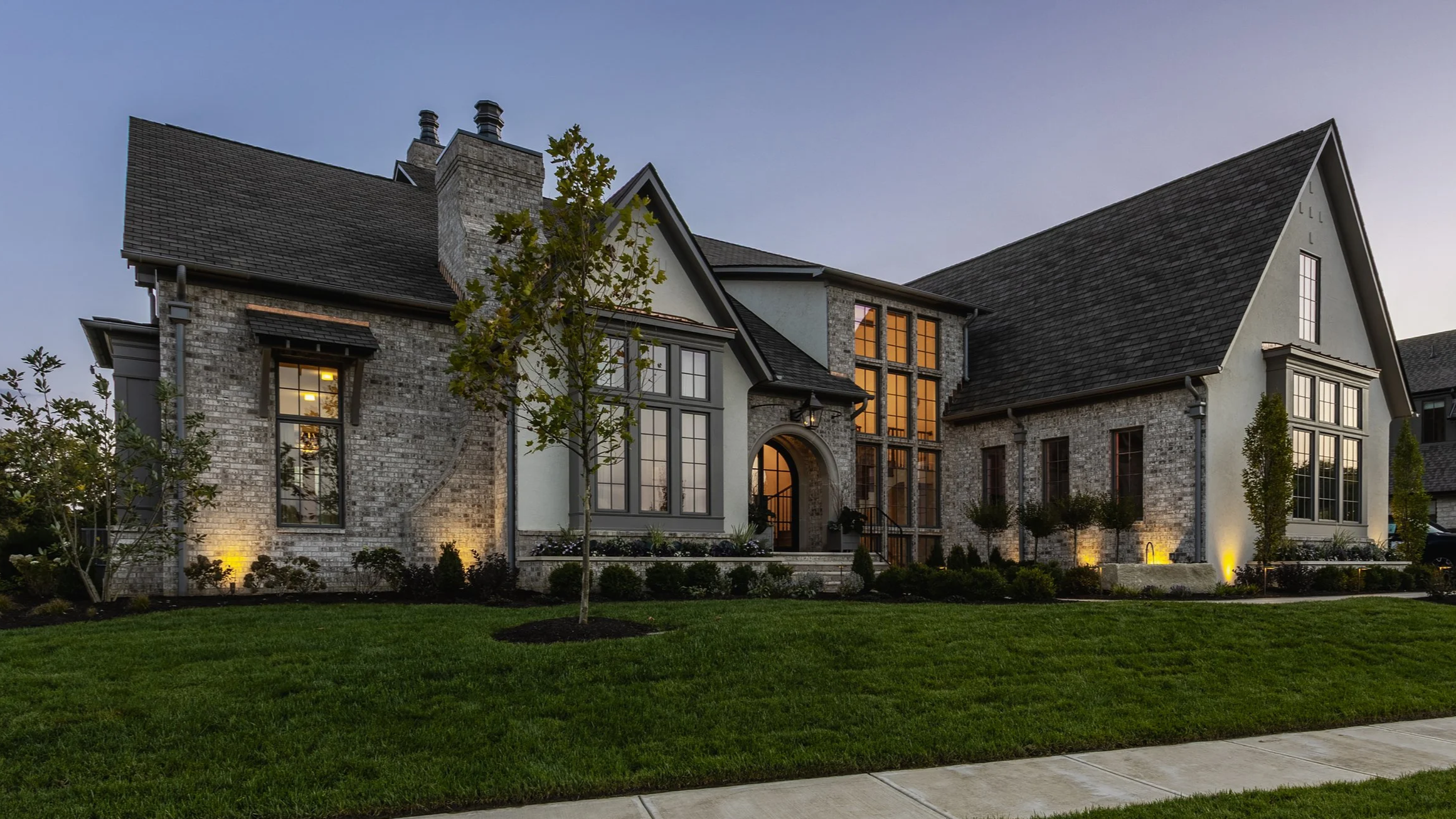 Front view of a large, modern house with stone and stucco exterior, multiple windows, and well-lit landscaping at dusk.