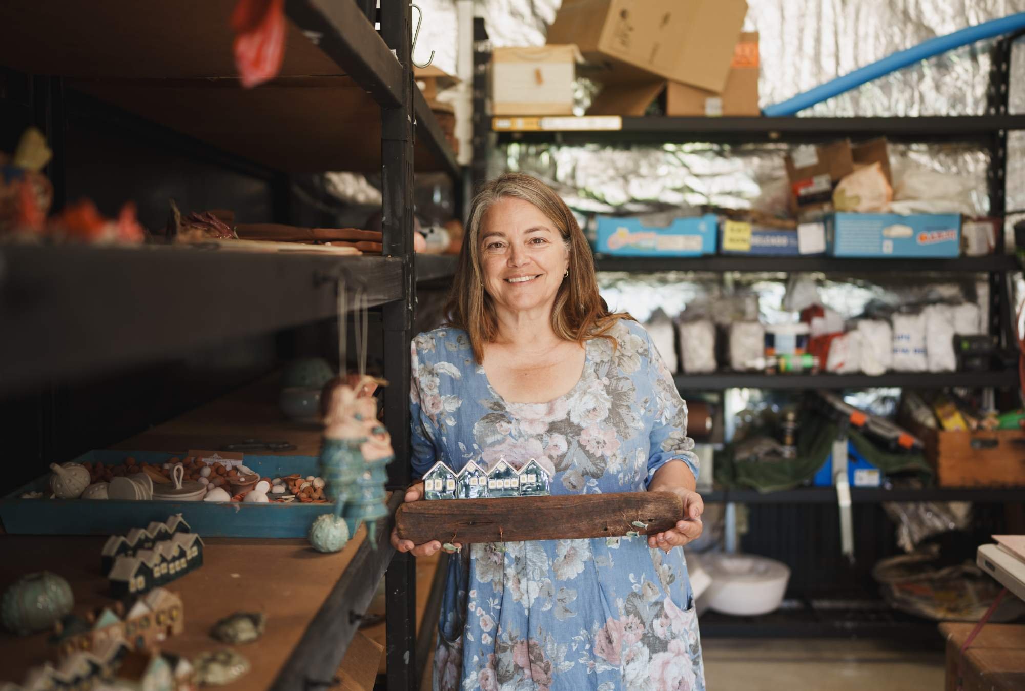 Smiling woman in floral dress holding a wooden piece at a craft or gift shop with shelves of various ceramic and décor items behind her.