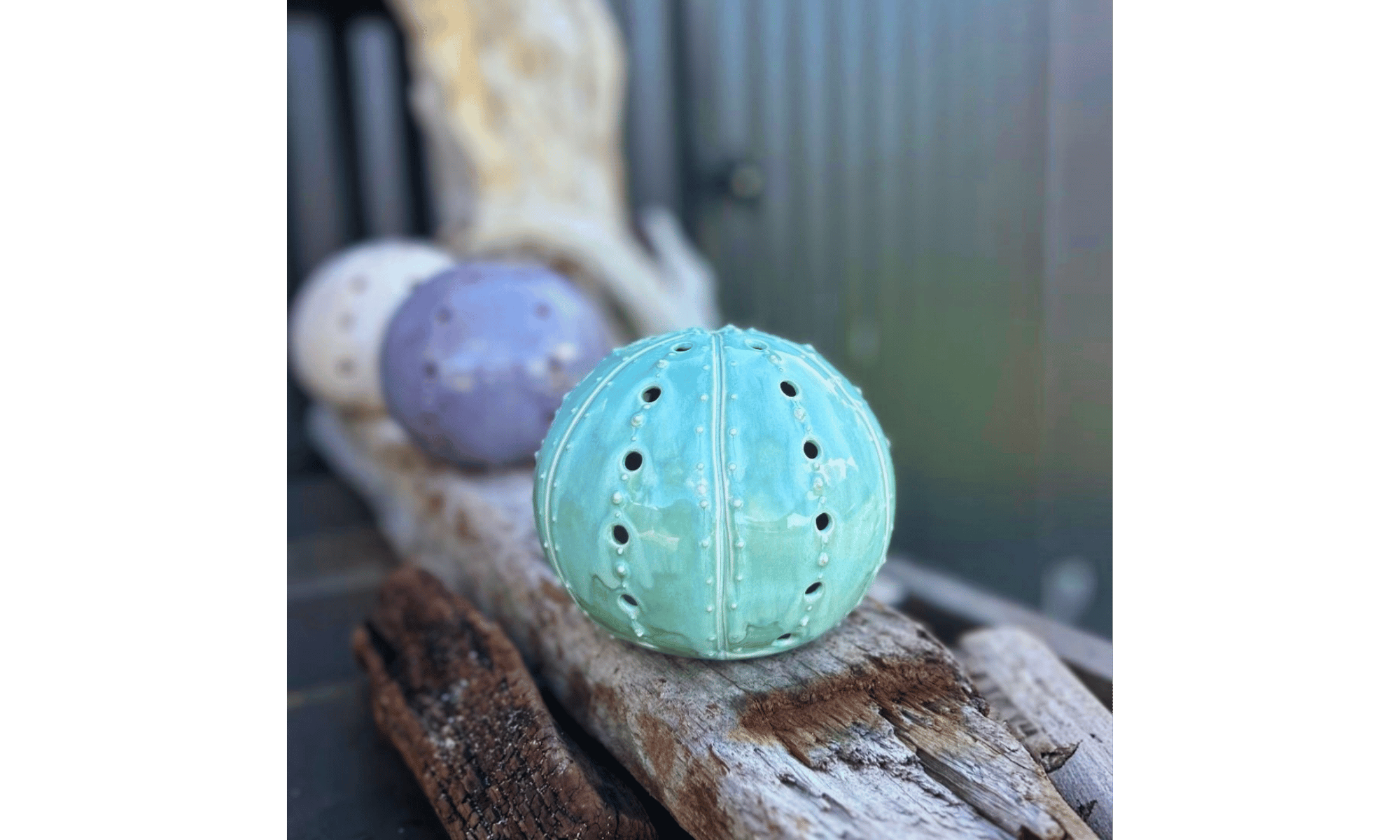 A light blue decorative ceramic ball with perforations, resting on piece of wood, with other similar decorative objects in the background.