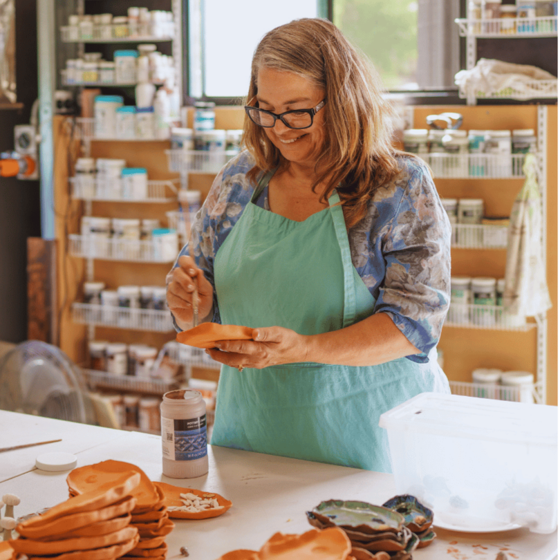 A woman with glasses and shoulder-length hair wearing an apron, smiling and working on a craft project in a workshop filled with shelves of supplies.