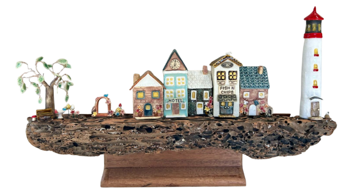 A miniature seaside village with a lighthouse, small houses, a tree, and decorative elements on a wooden base.