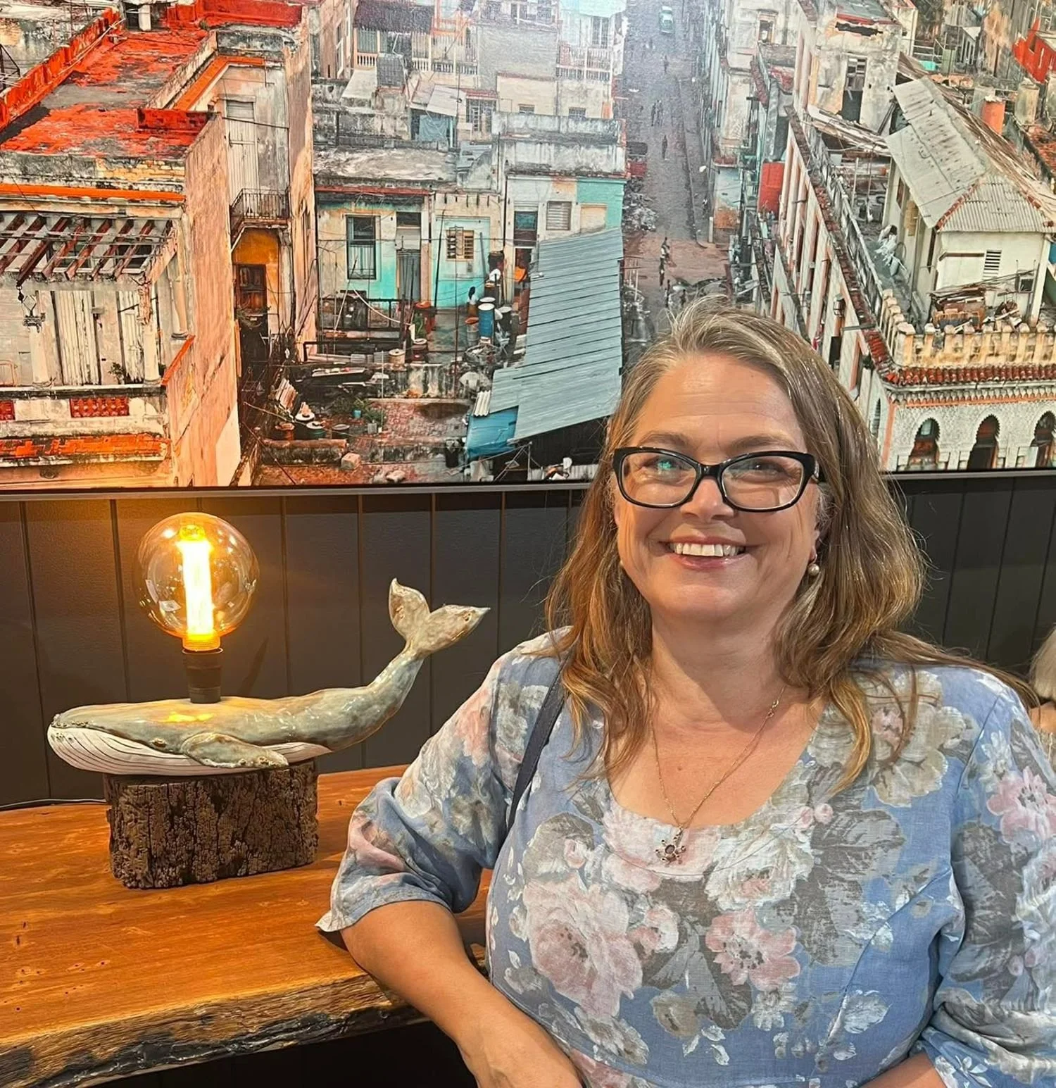 A woman with glasses and wavy hair smiling at a table with a whale-shaped lamp. Behind her, a wall features a cityscape view of old buildings with weathered facades and rooftops. The whale lamp has an exposed lightbulb and is mounted on a wooden base.