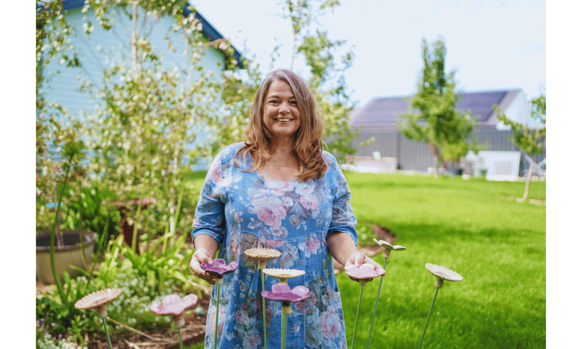 Woman in a floral dress standing in a garden with flowers, holding gardening tools. She is smiling, with a blue house and trees in the background.