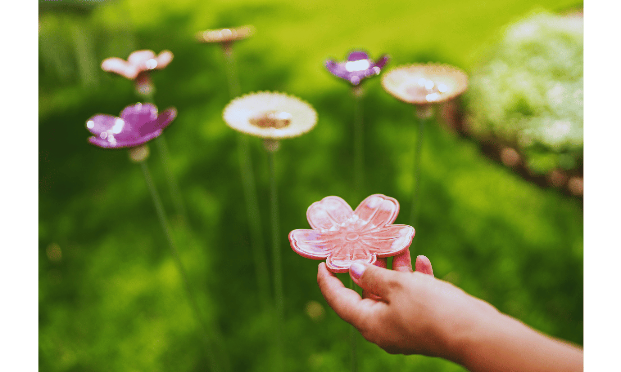 A person's hand holding a pink glass flower-shaped object in front of a blurred background of colorful glass flowers and green foliage.