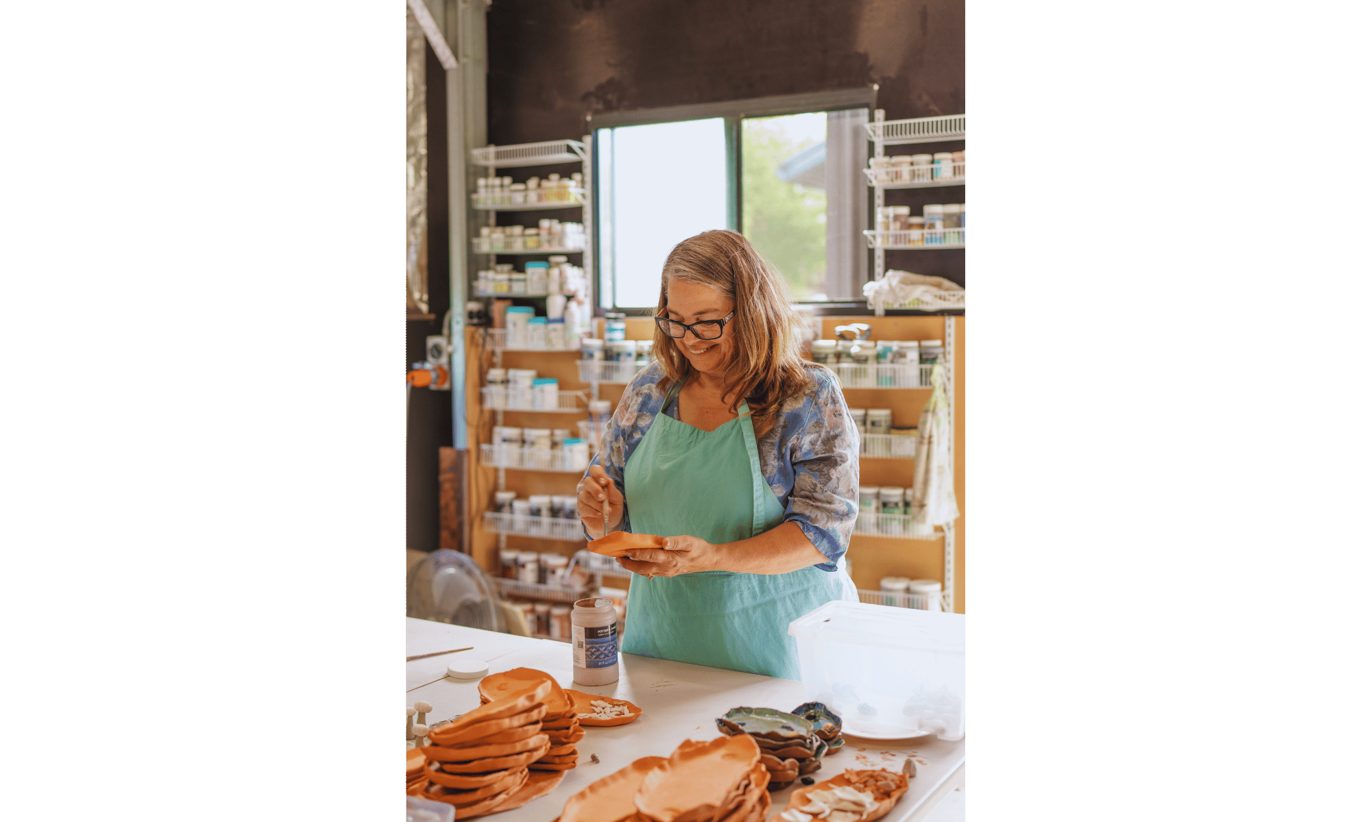 A woman wearing glasses and a green apron, smiling while working with clay or ceramic pieces in a craft room or studio, with shelves of supplies and a window in the background.