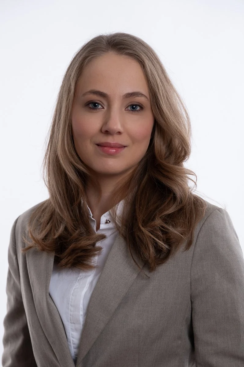 A portrait of a young woman with long, wavy light brown hair, wearing a beige blazer and white shirt, against a white background.