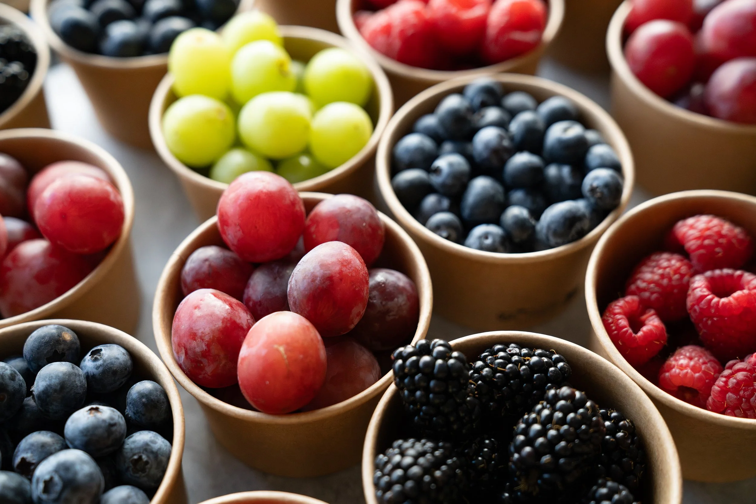 Assorted fresh berries in small paper cups, including blueberries, blackberries, red raspberries, green grapes, and red grapes.