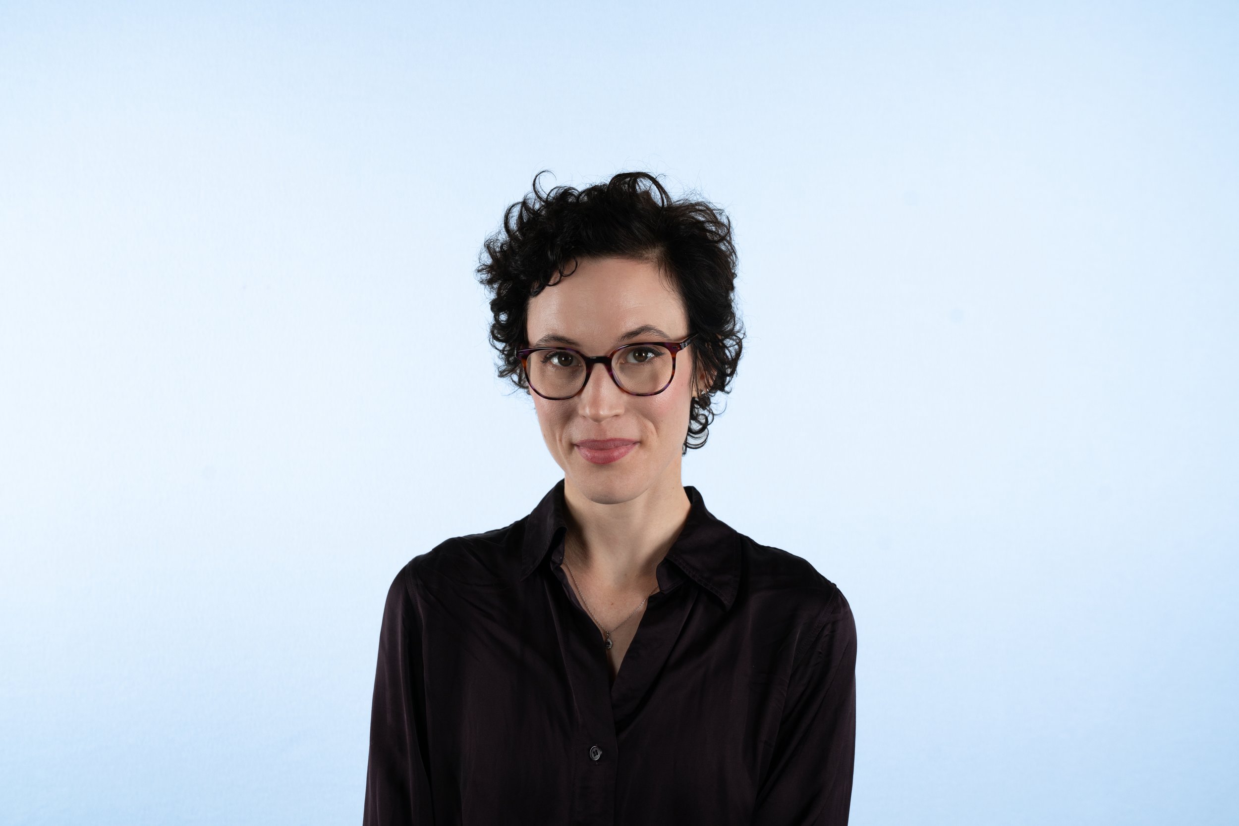 A woman with short curly dark hair, wearing glasses and a black button-up shirt, standing against a plain light blue background.