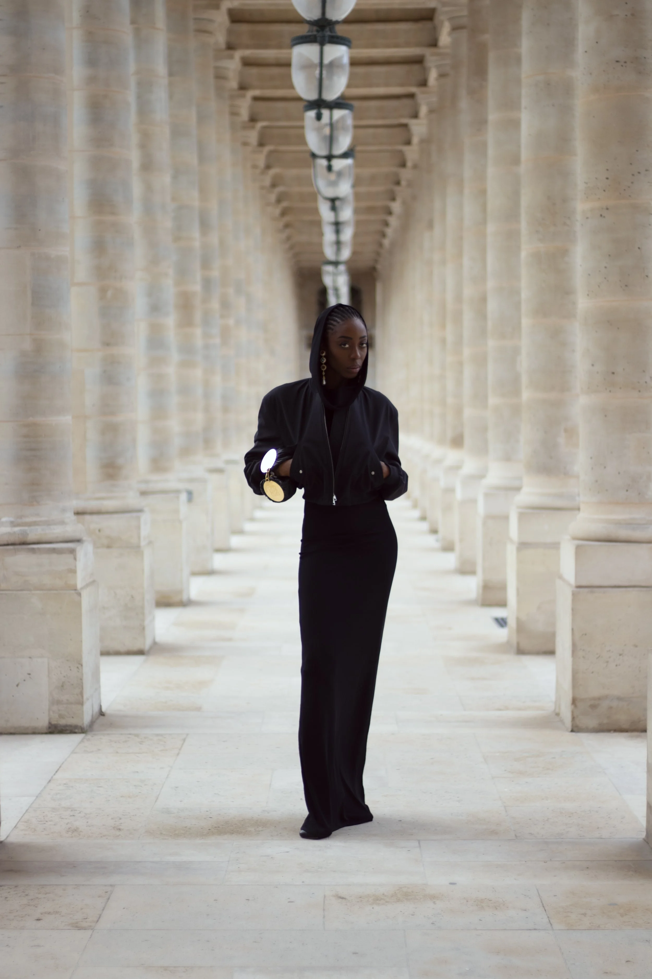 A woman wearing a black hoodie, long black skirt, and earrings stands in a columned corridor with beige stone walls and floor, lined with overhead lamps.