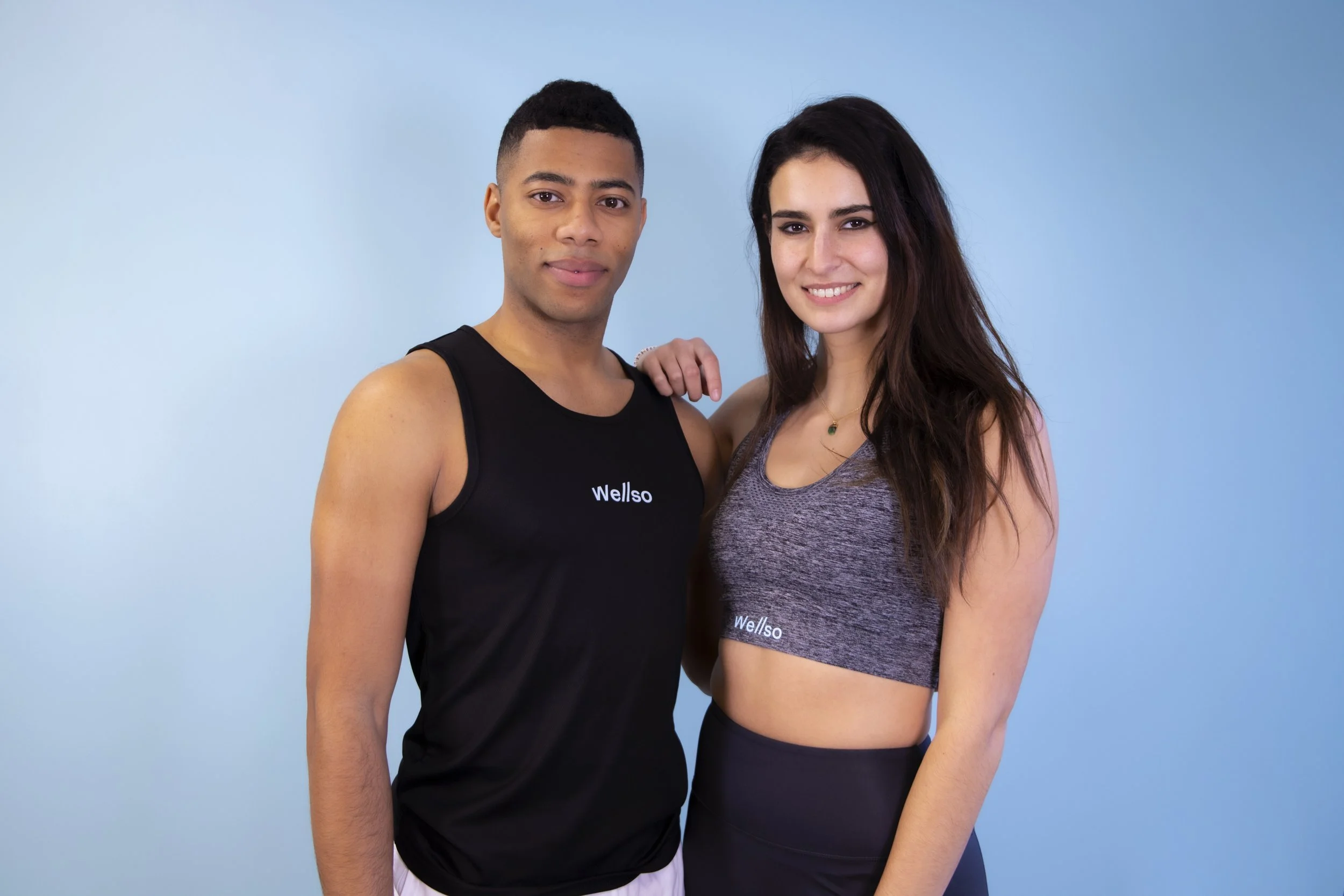 A young man and woman standing close together against a blue background, smiling at the camera. The man is wearing a black tank top with the word 'Wellso' on it, and the woman is wearing a gray athletic tank top with the same 'Wellso' logo.
