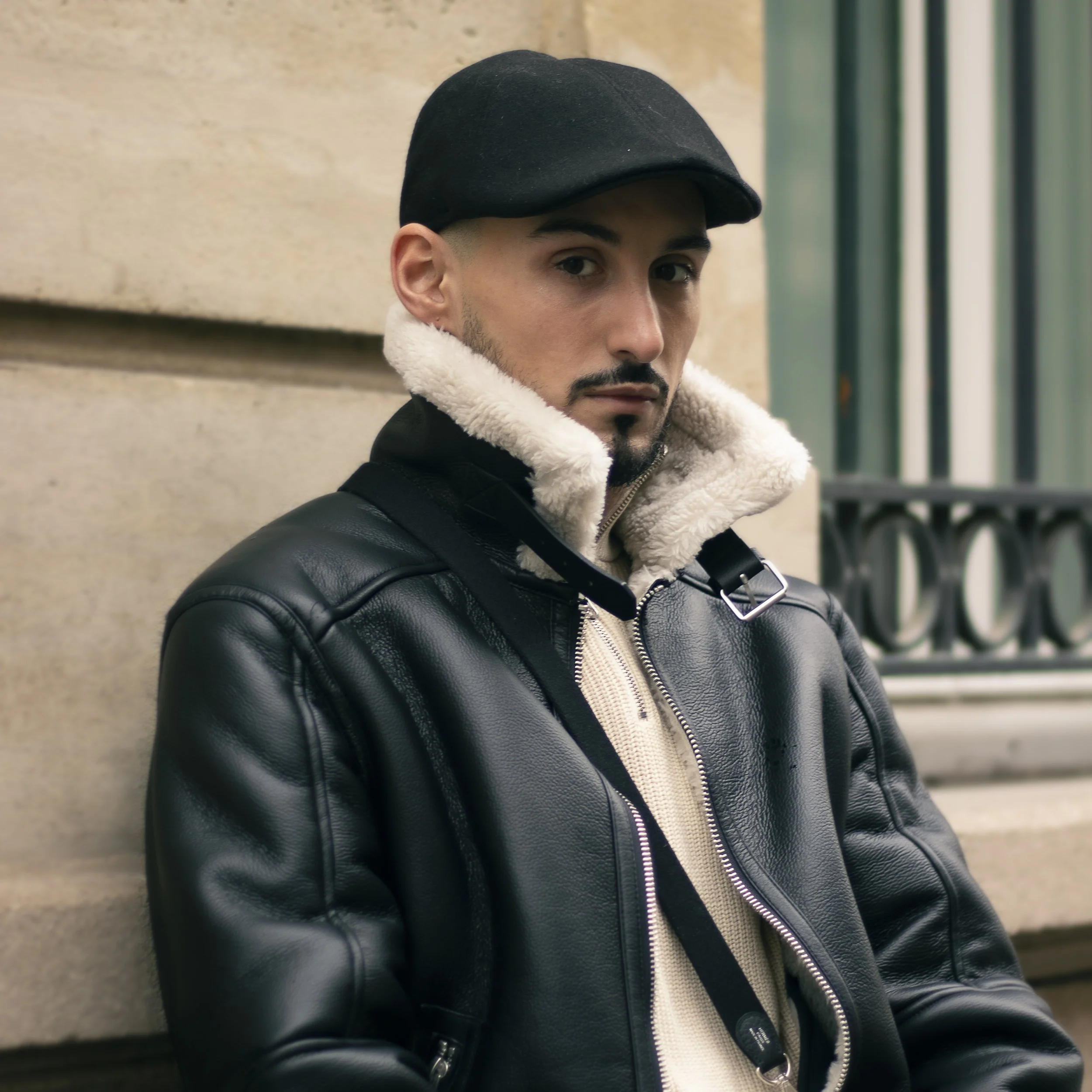 A young man with dark hair and a mustache wearing a black cap, a black leather jacket, and a cream-colored hoodie, leaning against a stone wall with green metal bars in the background.