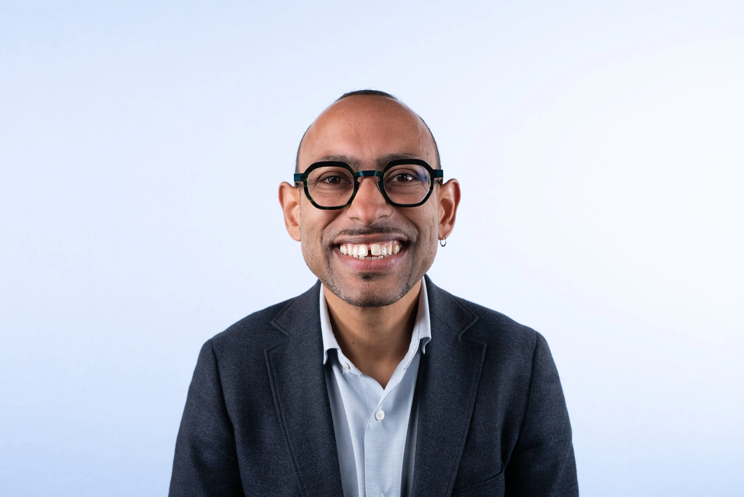 A man with glasses, a beard, and a earring smiling in a suit jacket and white shirt against a light blue background.