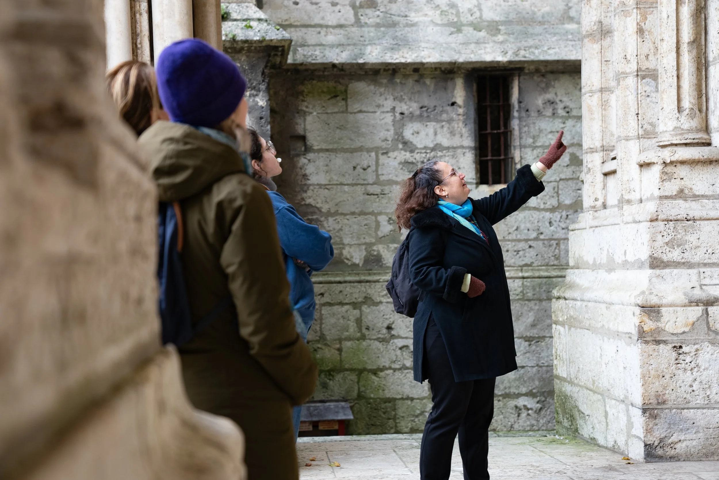 A group of people on a tour, listening to a woman guide pointing at the wall of an old stone building.
