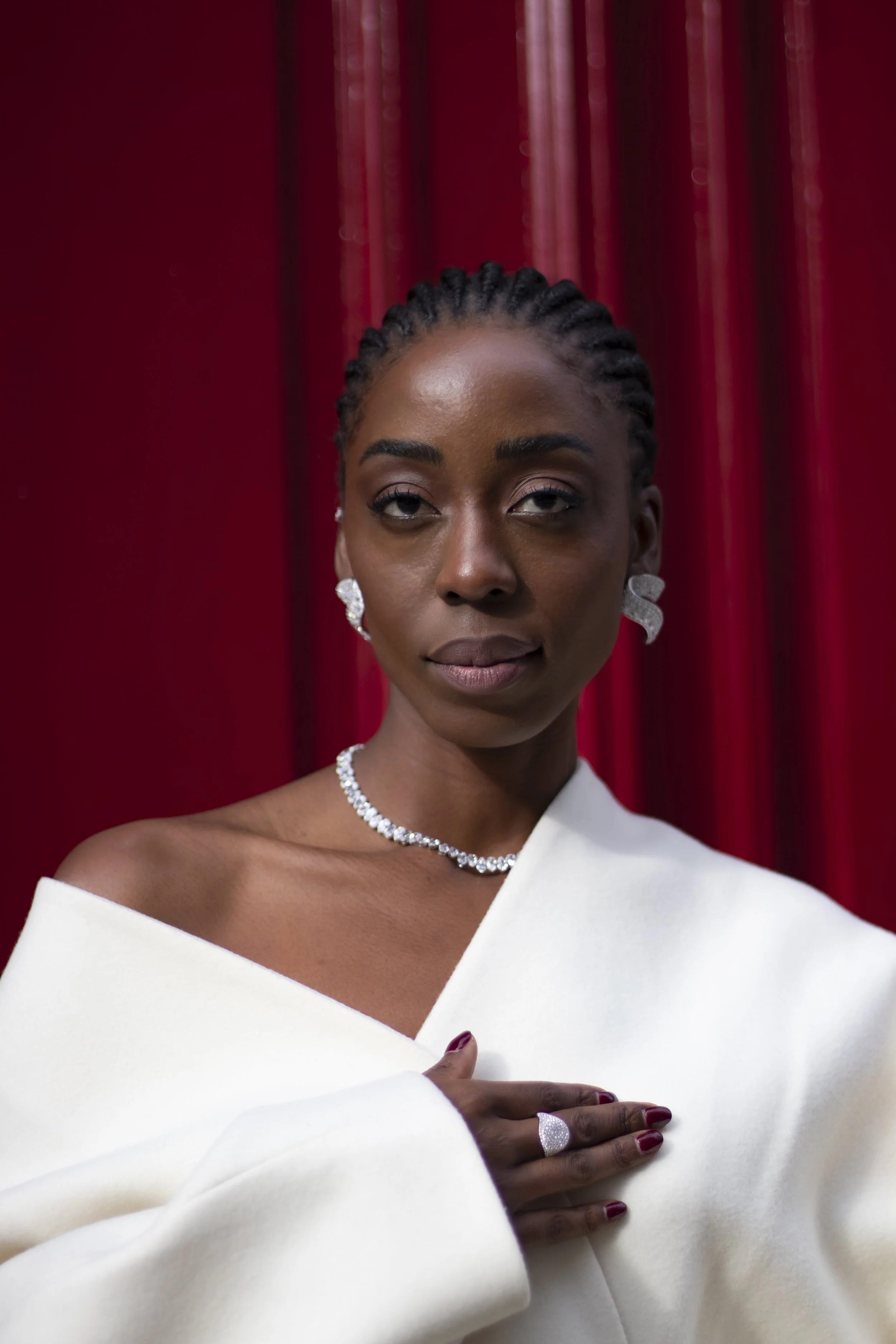 A woman dressed in an off-the-shoulder white gown, with earrings and a necklace, poses against a red curtain backdrop. Her right hand is placed over her chest, revealing a shiny ring and matching nails.