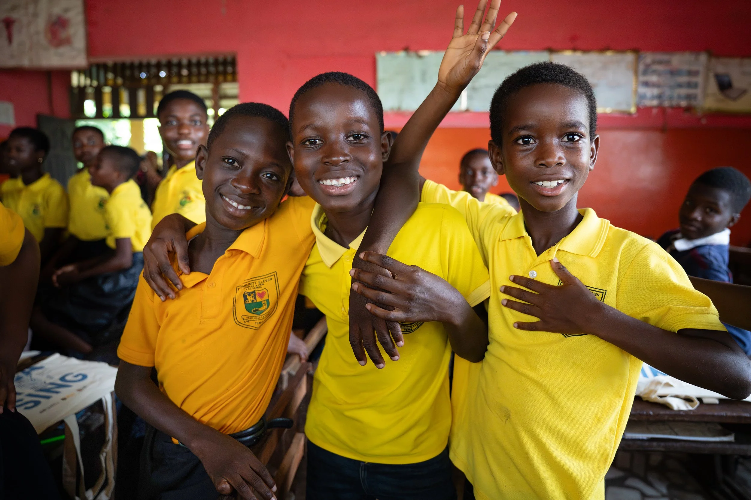 Smiling children in yellow shirts standing together in a classroom, with some raising their hands and one child hugging another.