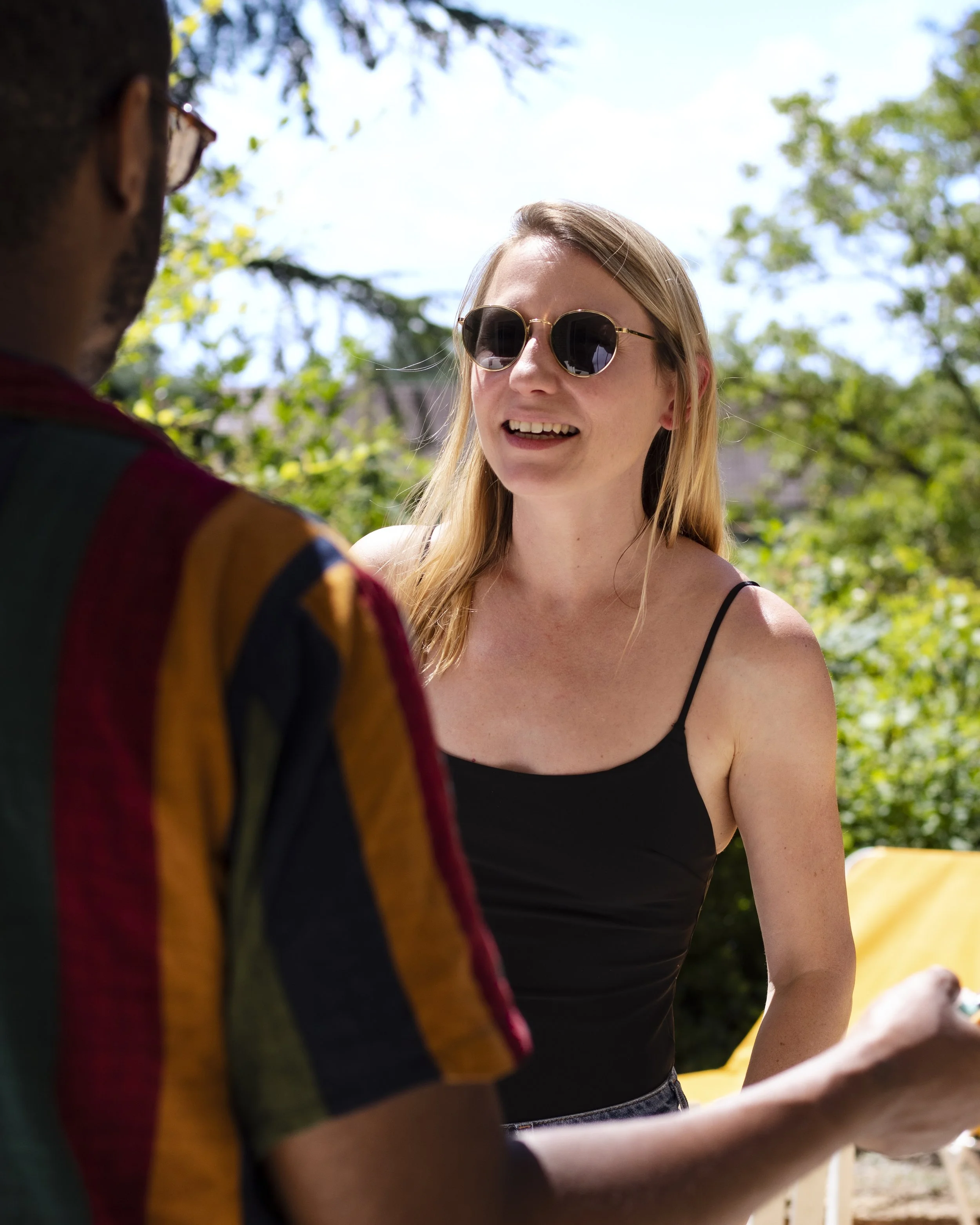 A woman with long blonde hair and wearing black sunglasses is smiling and talking to a man whose face isn't visible. They are outdoors with trees and blue sky in the background.
