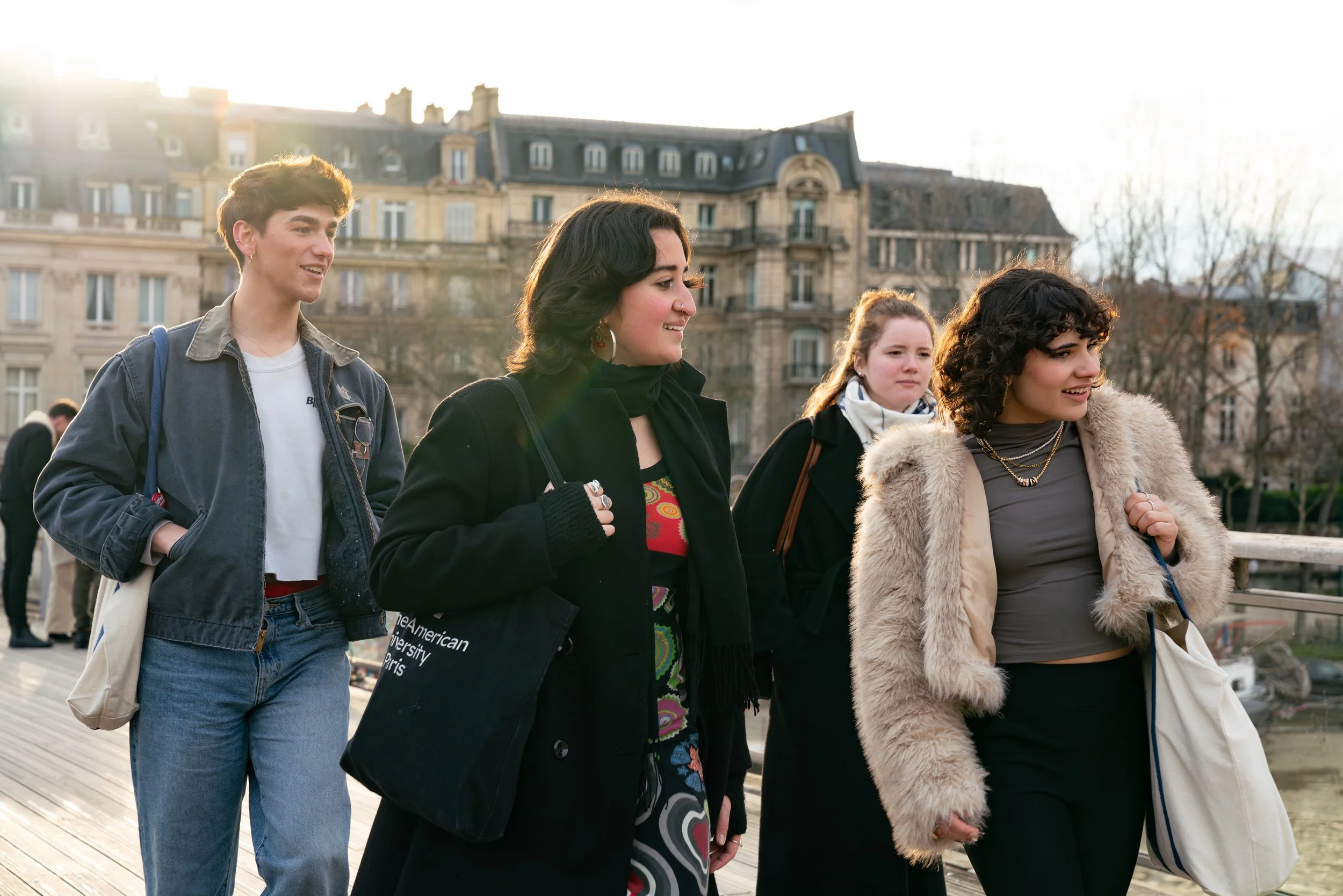 A group of four young women walking outdoors on a sunny day with historic buildings in the background.