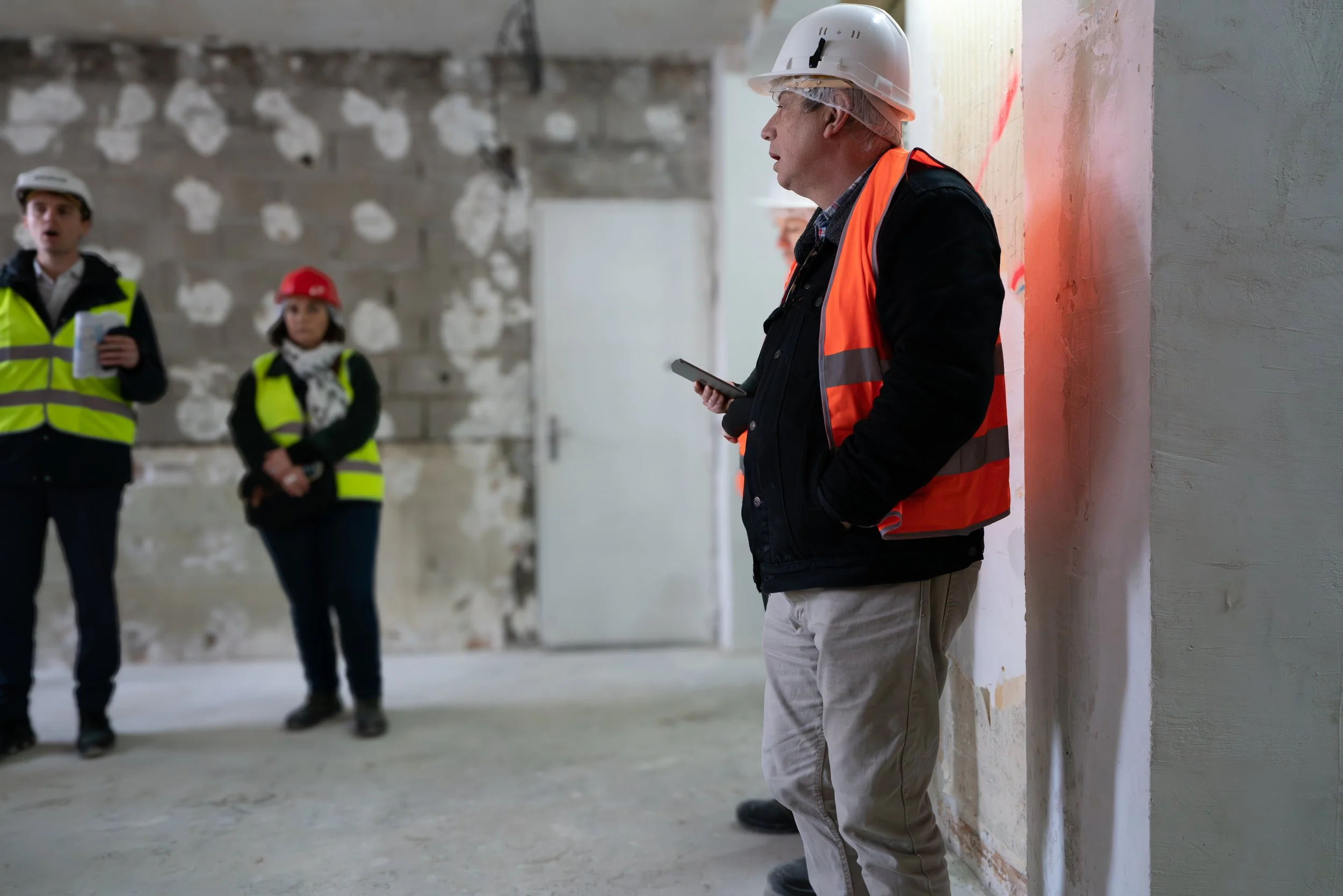 A construction worker leans against a wall while wearing a white hard hat, orange safety vest, and casual clothing at an indoor construction site. Two other individuals in yellow safety vests and hard hats are seen in the background, one holding a no