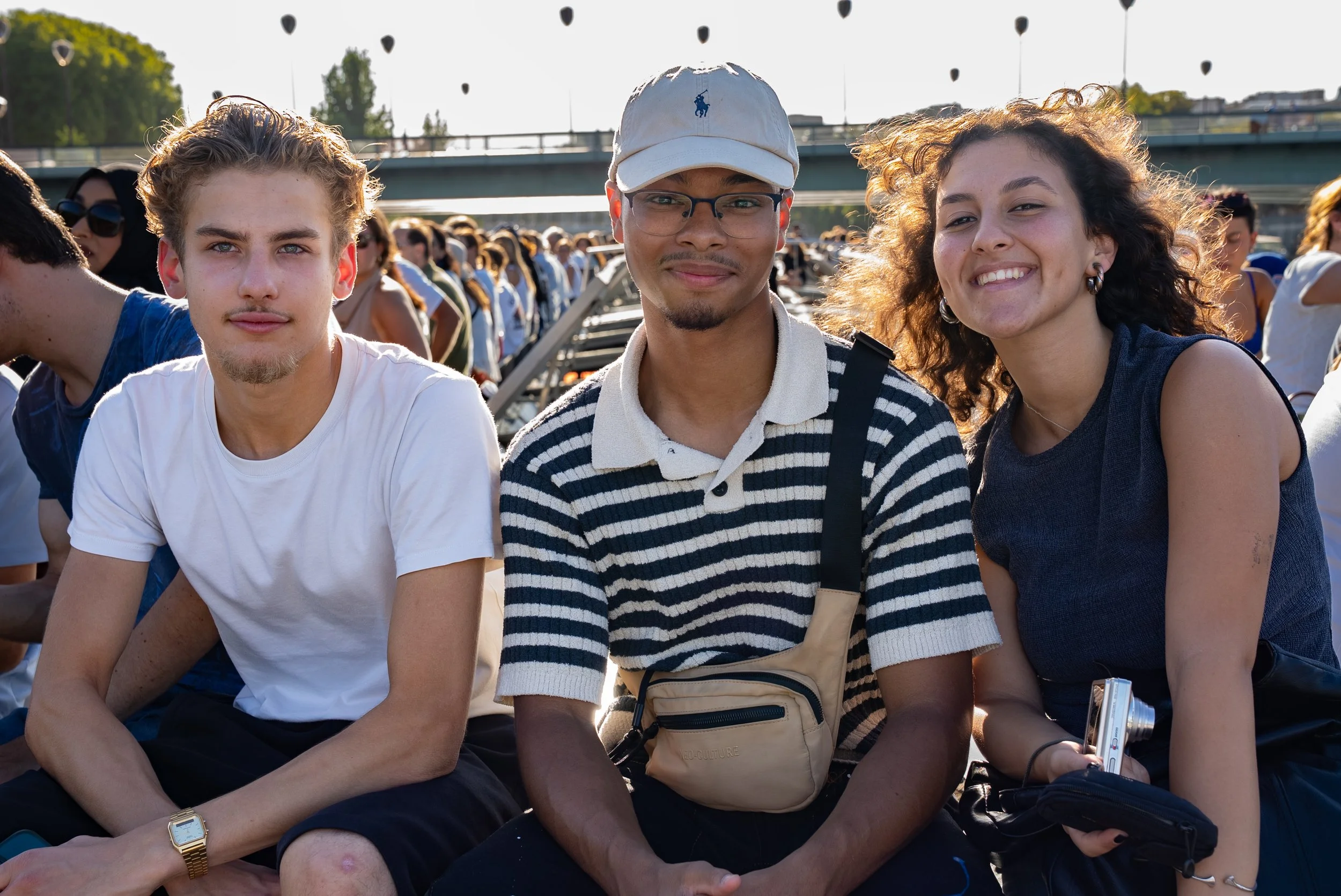 Three young people sitting outdoors at event, smiling at camera with crowd in background.