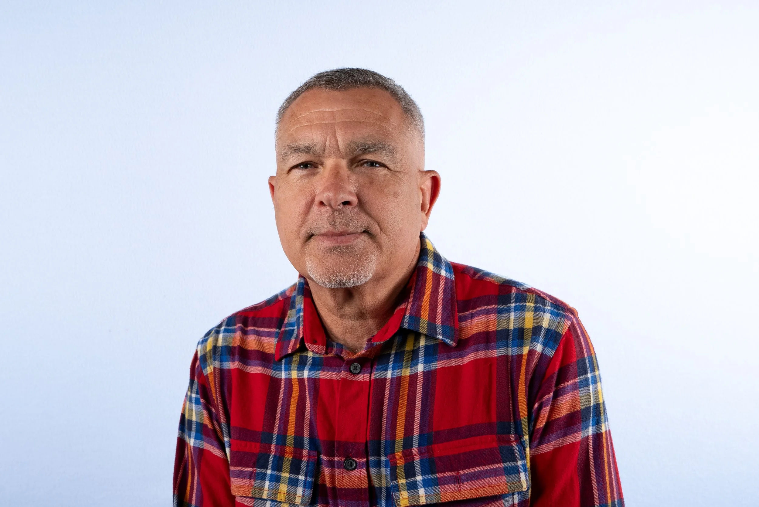 A middle-aged man with short grey hair, a mustache, and a goatee is wearing a red, blue, yellow, and green plaid shirt, standing in front of a white background.