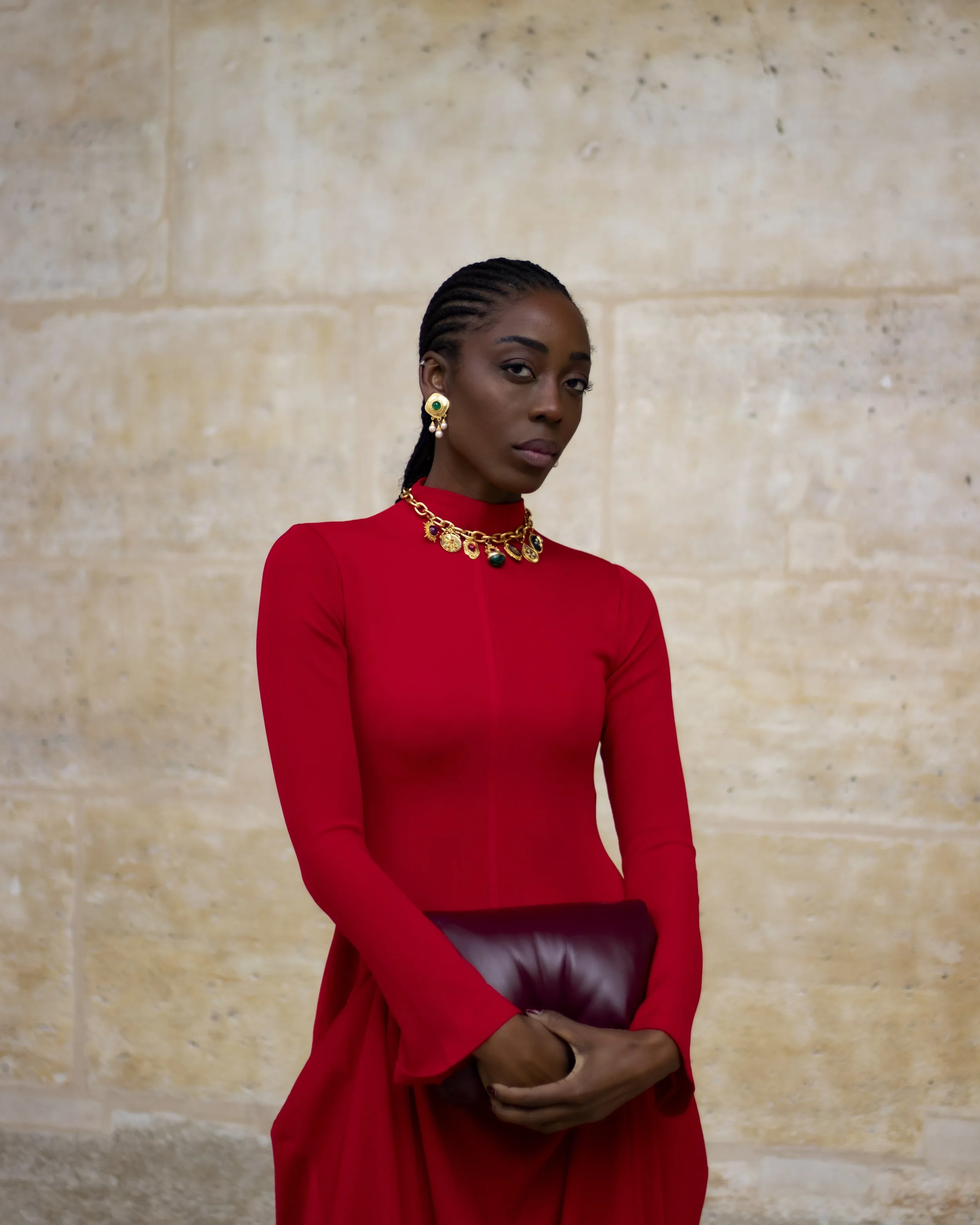 A woman with dark skin, braided hair, and serious expression, dressed in a red top, gold jewelry with green accents, holding a brown clutch purse, standing against a beige stone wall.
