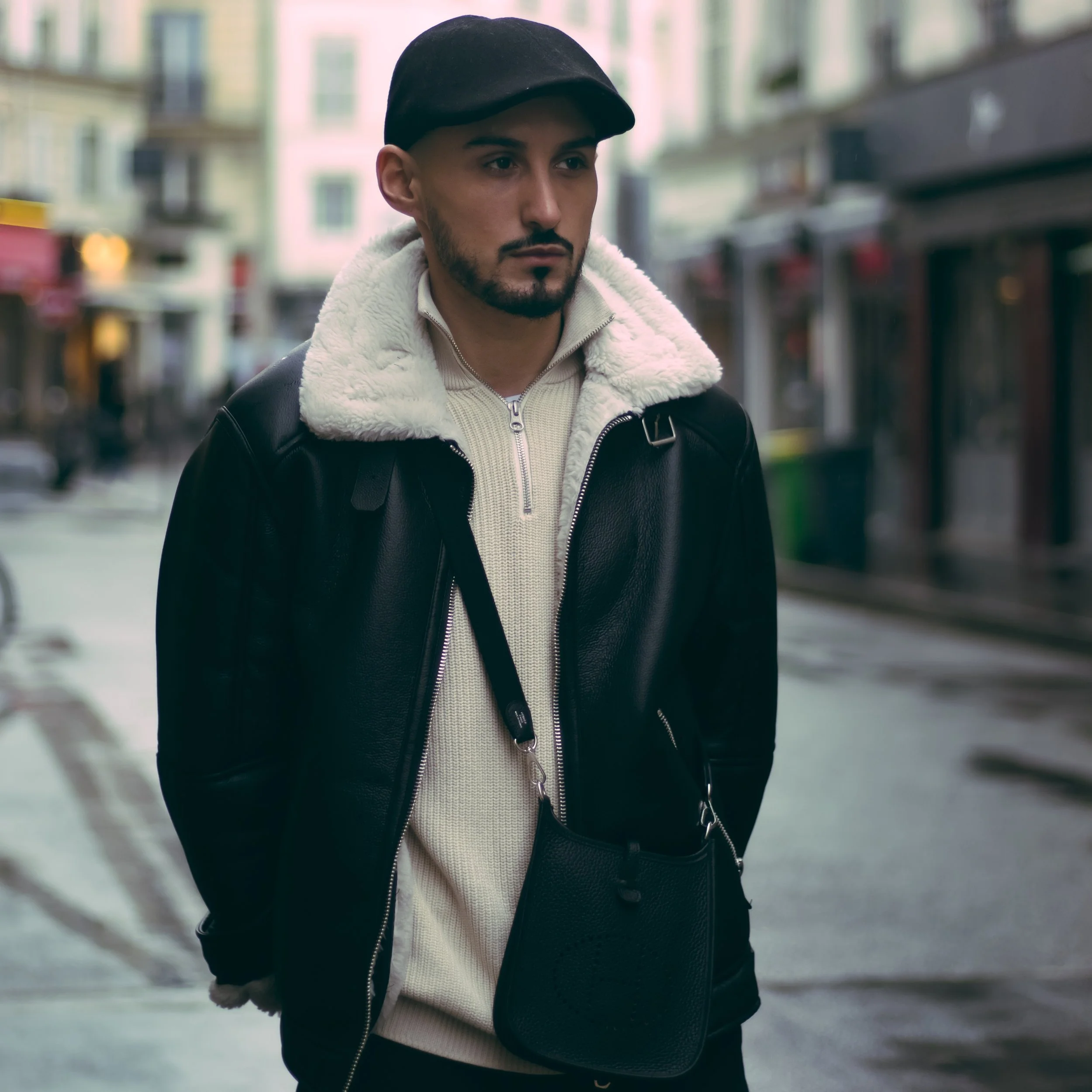 A young man with a beard wears a black cap, a black leather jacket with a fluffy white collar, and a cream-colored sweater underneath. He walks on a city street with blurred buildings in the background, looking serious and focused.
