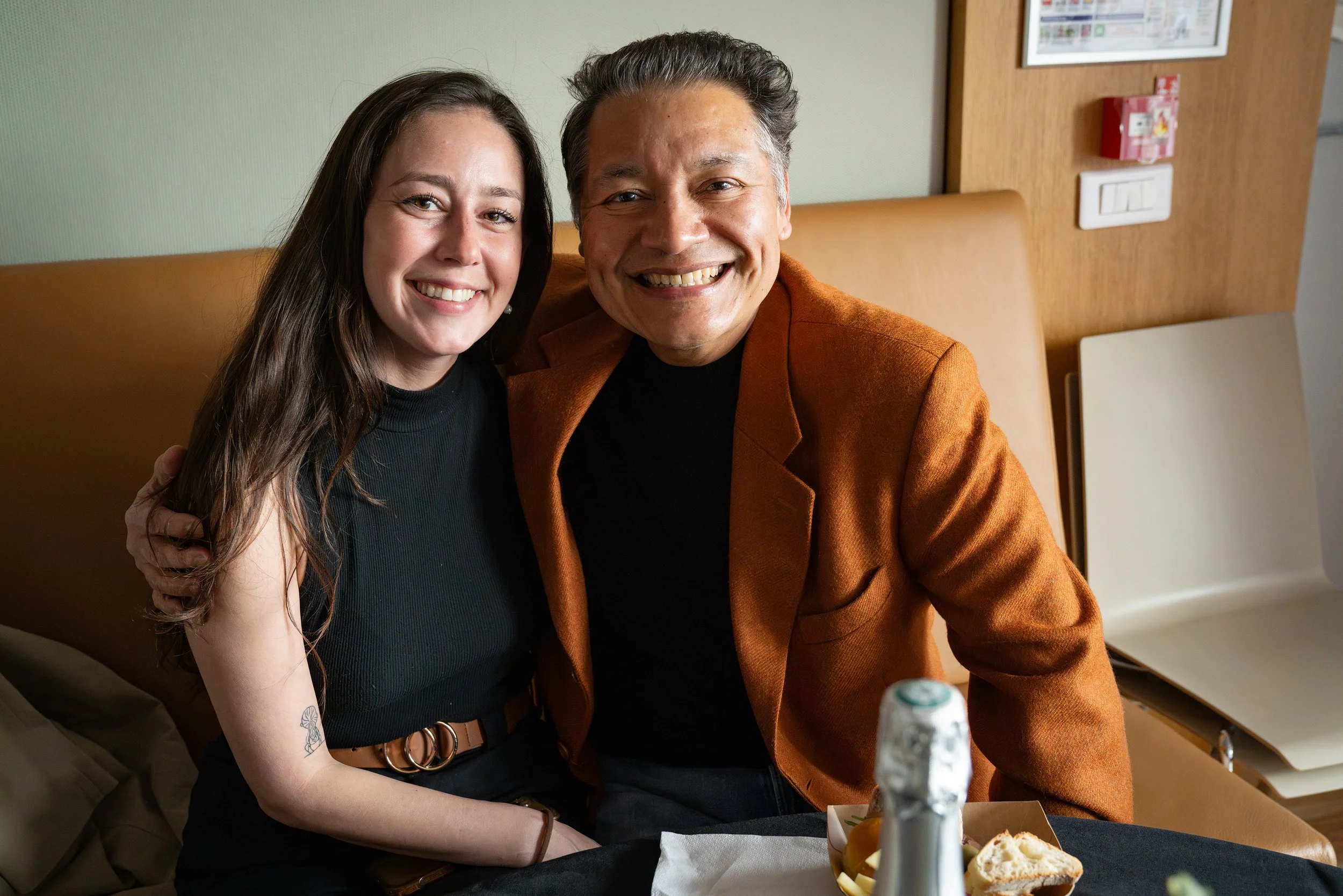 A young woman and an older man smiling, sitting close together at a restaurant table with food and a bottle in view.