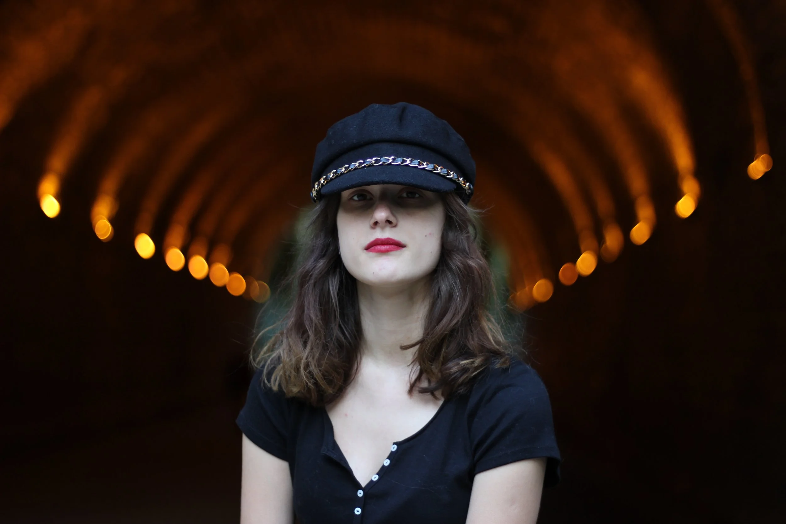 A young woman with long brown hair wearing a black hat with a metallic chain band, standing in front of a tunnel illuminated with warm orange lights, looking directly at the camera.