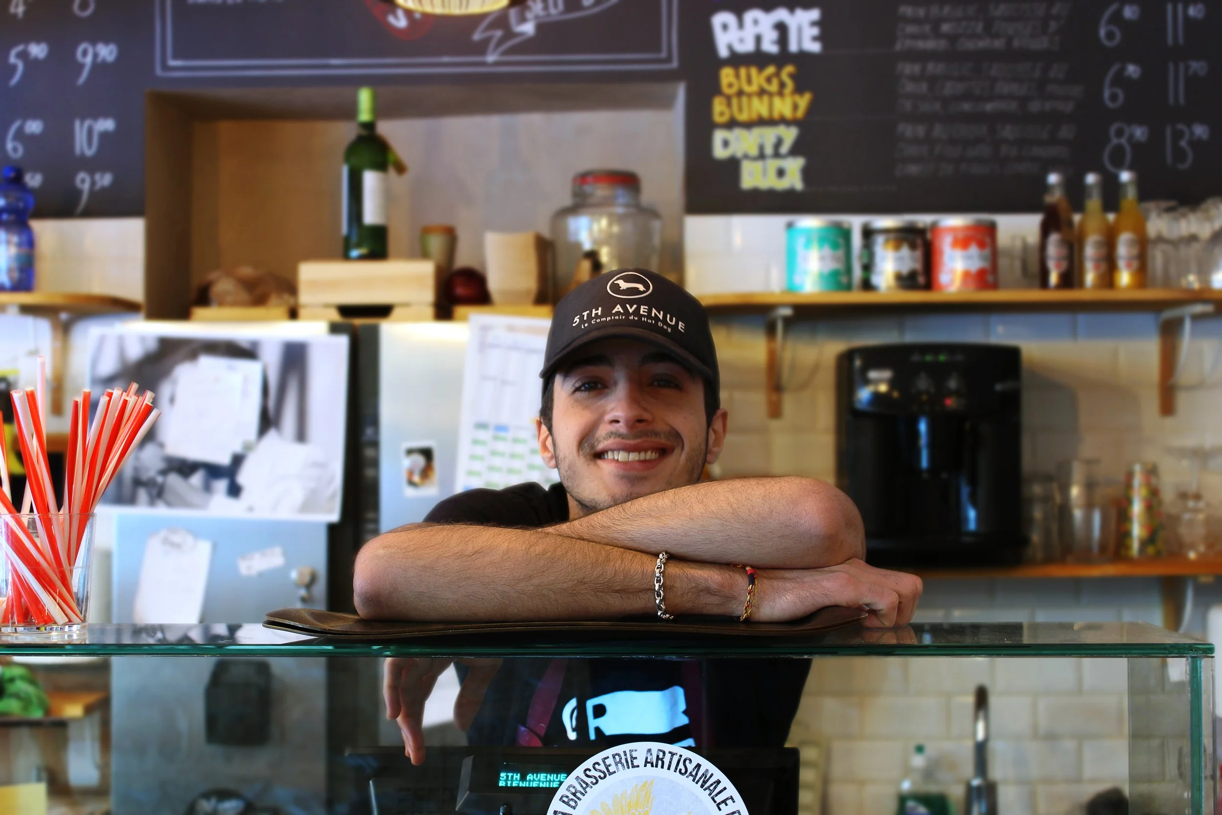 A man with a smiling expression resting his head and arms on a glass counter in a cafe or restaurant. He is wearing a black cap and a bracelet. In the background, there is a menu board with colorful text, and various condiments and drink bottles are 