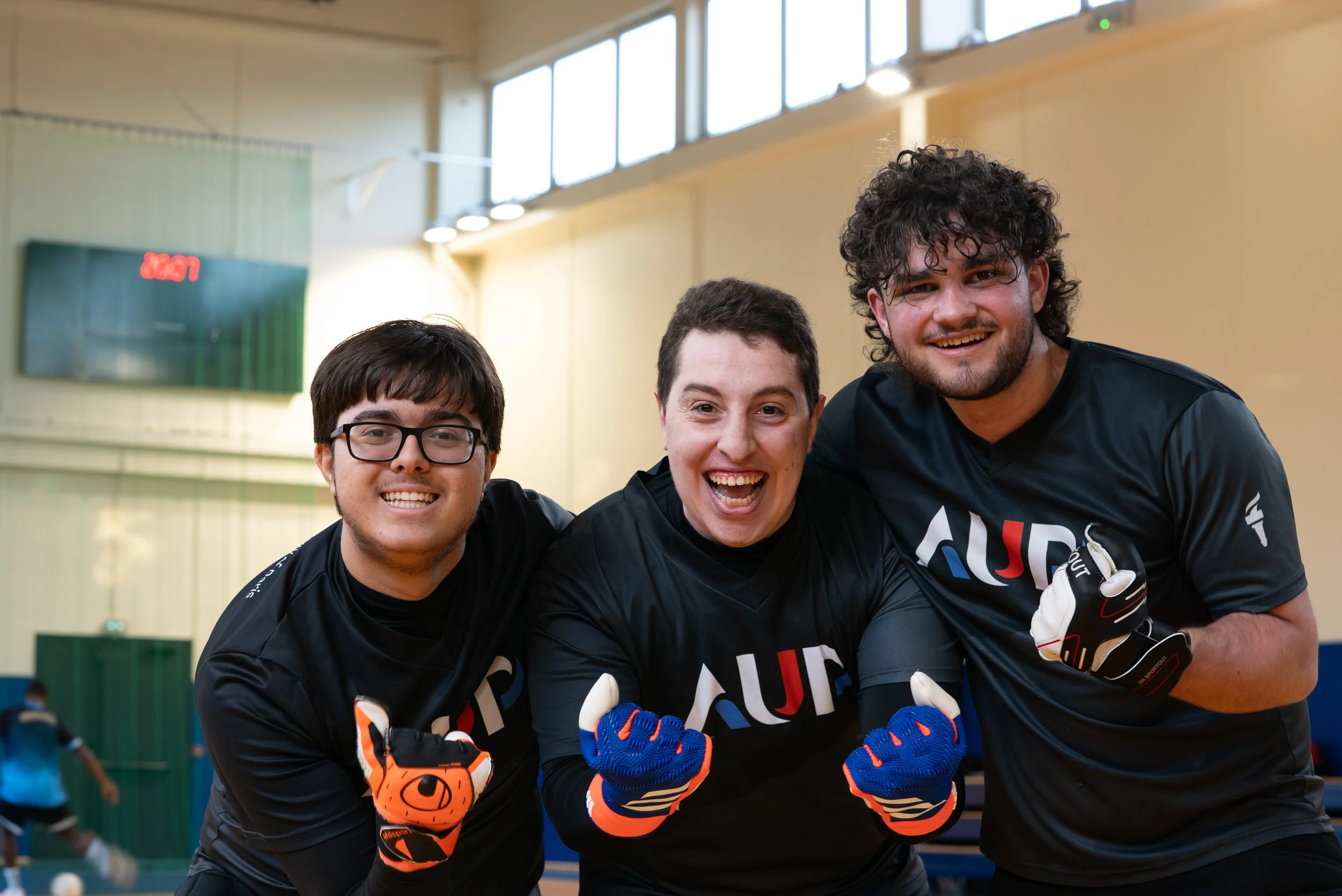 Three young men in black sports jerseys smiling and posing together indoors, with one wearing goalkeeper gloves, in what appears to be a gym or sports hall.