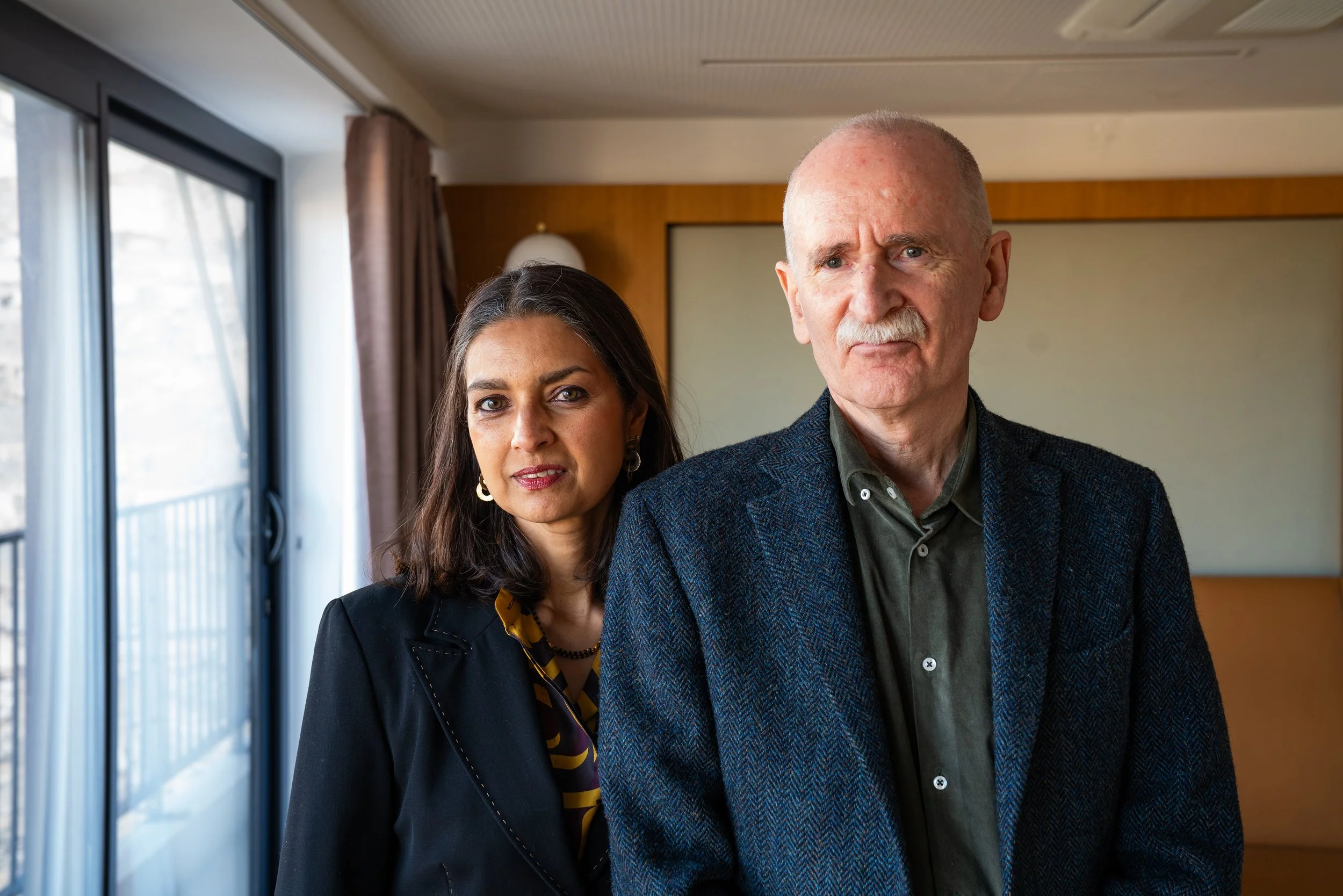 A man and a woman posing together indoors with a sliding glass door and curtains in the background.