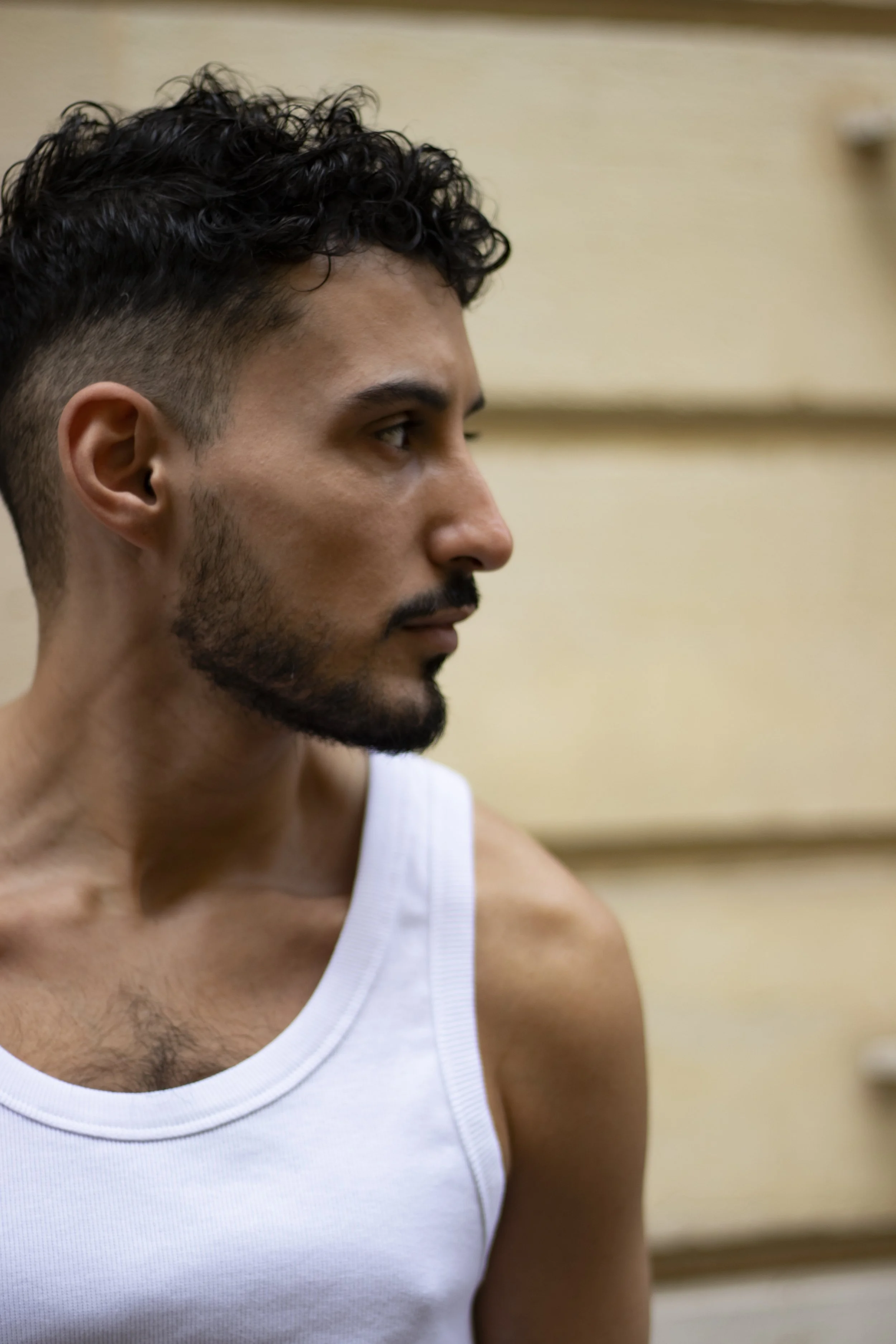 Profile of a young man with curly black hair, a beard, wearing a white sleeveless shirt, looking to the right with a neutral expression, against a blurred indoor background.