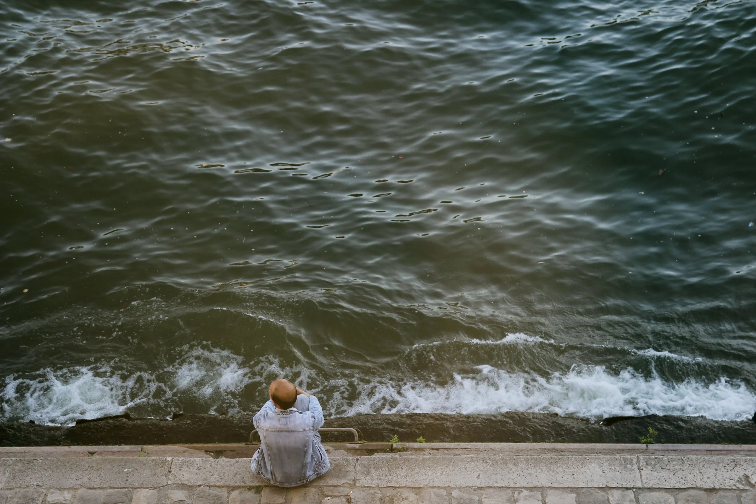 A person in a gray jacket sitting on a stone ledge facing the water, which has small waves and ripples. The person appears to be gazing at the water.