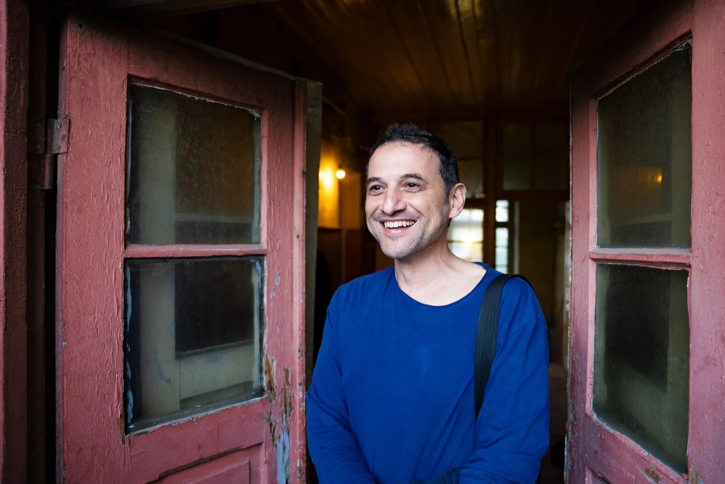 A man with short dark hair and a big smile, wearing a blue shirt, standing at a doorway with red wooden doors and glass panes, inside a cozy wooden interior with warm lighting.