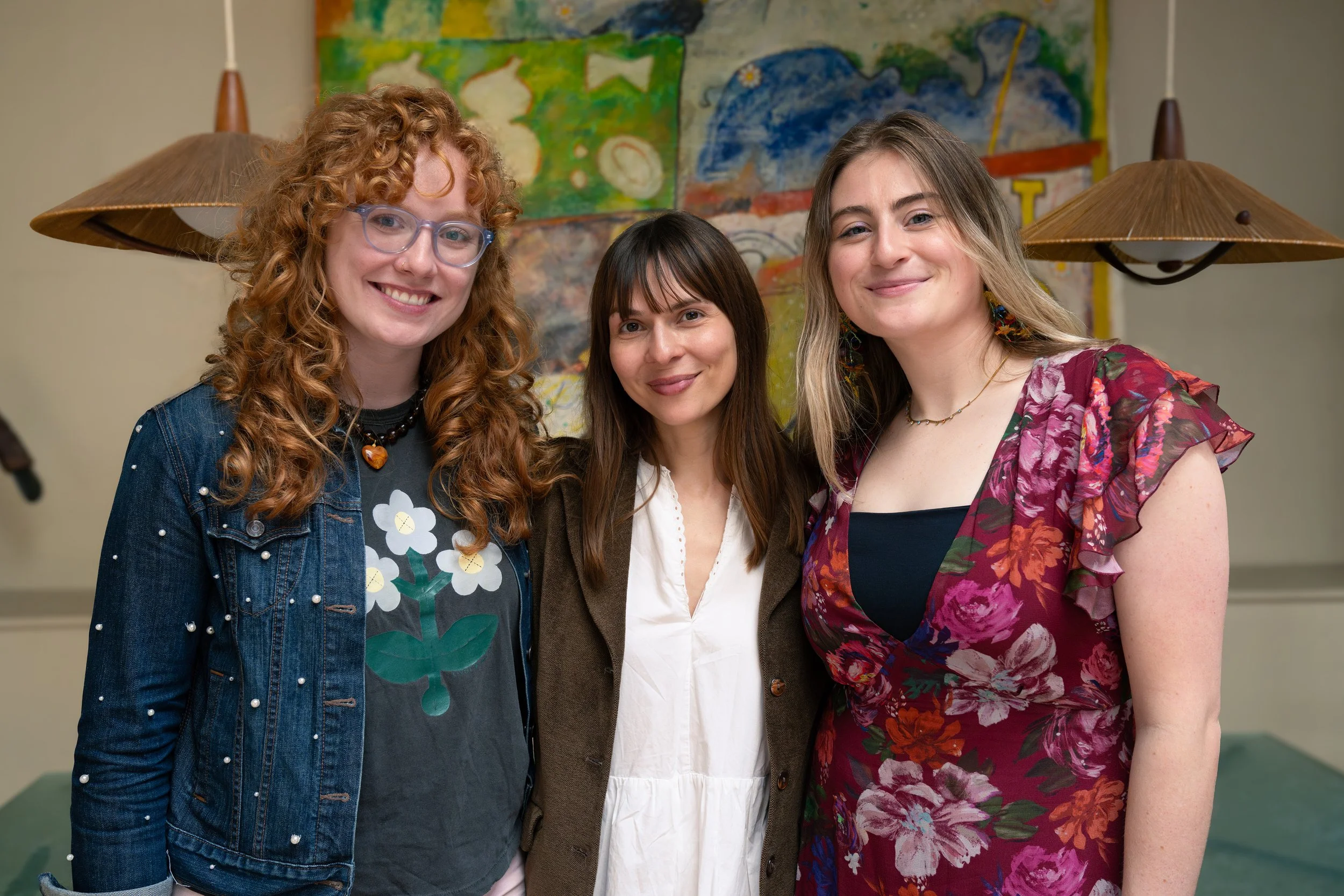 Three women smiling and standing close together indoors, with a colorful abstract painting and two hanging lamps in the background.