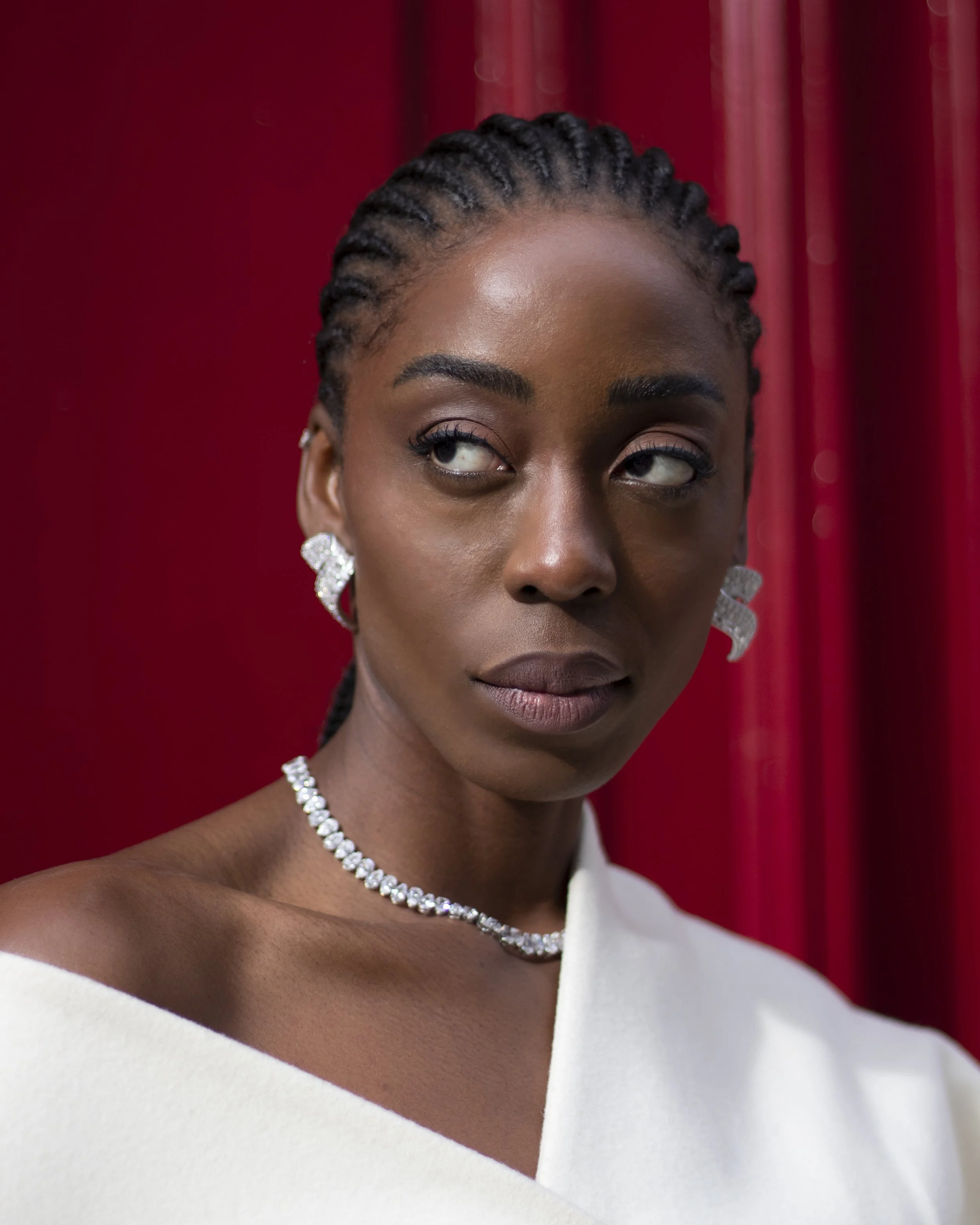 Close-up of a woman with braided hair, wearing diamond earrings and a diamond necklace, with a red curtain in the background.