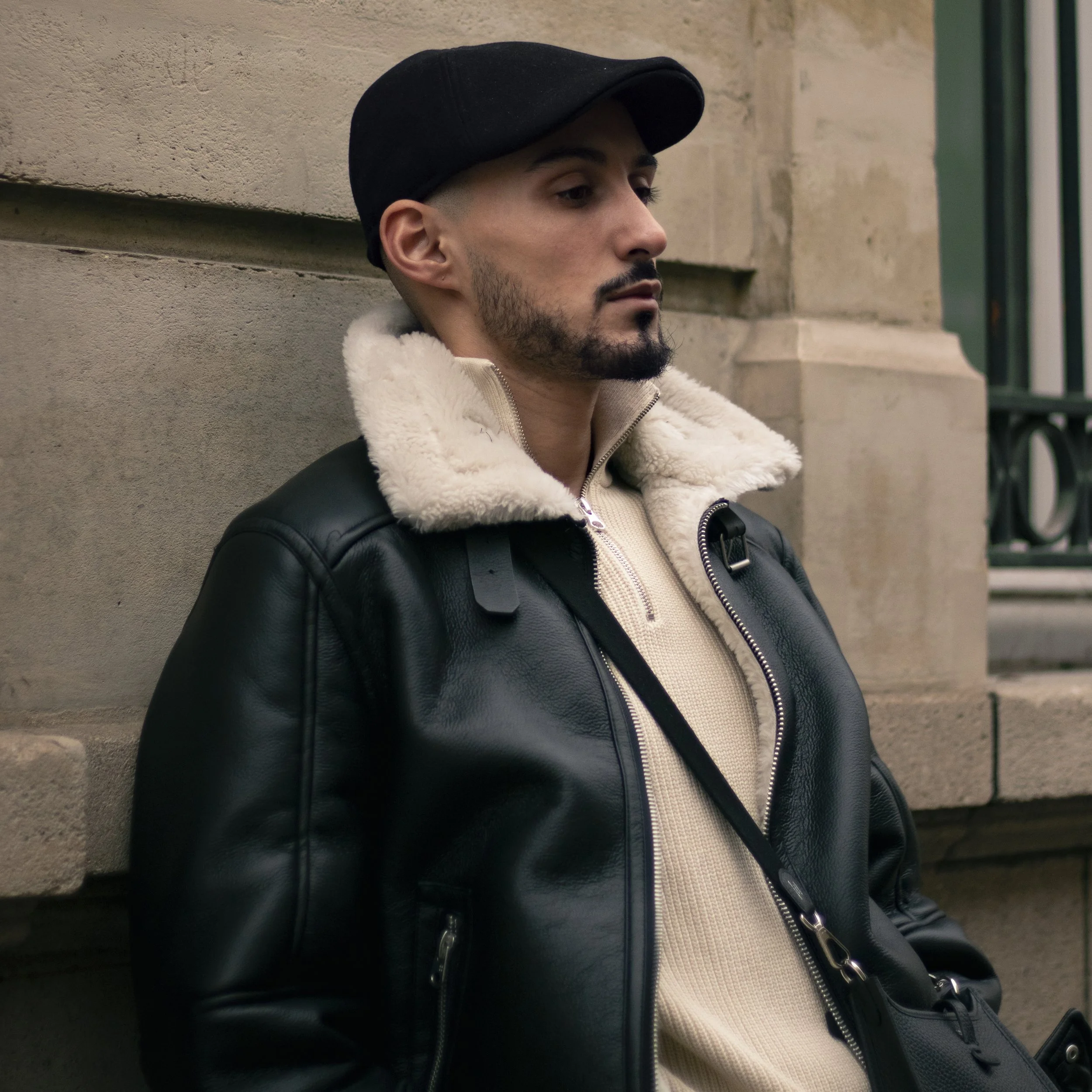 A young man with a beard and mustache wearing a black cap, cream-colored sweater, and black leather jacket, sitting against a beige brick wall.