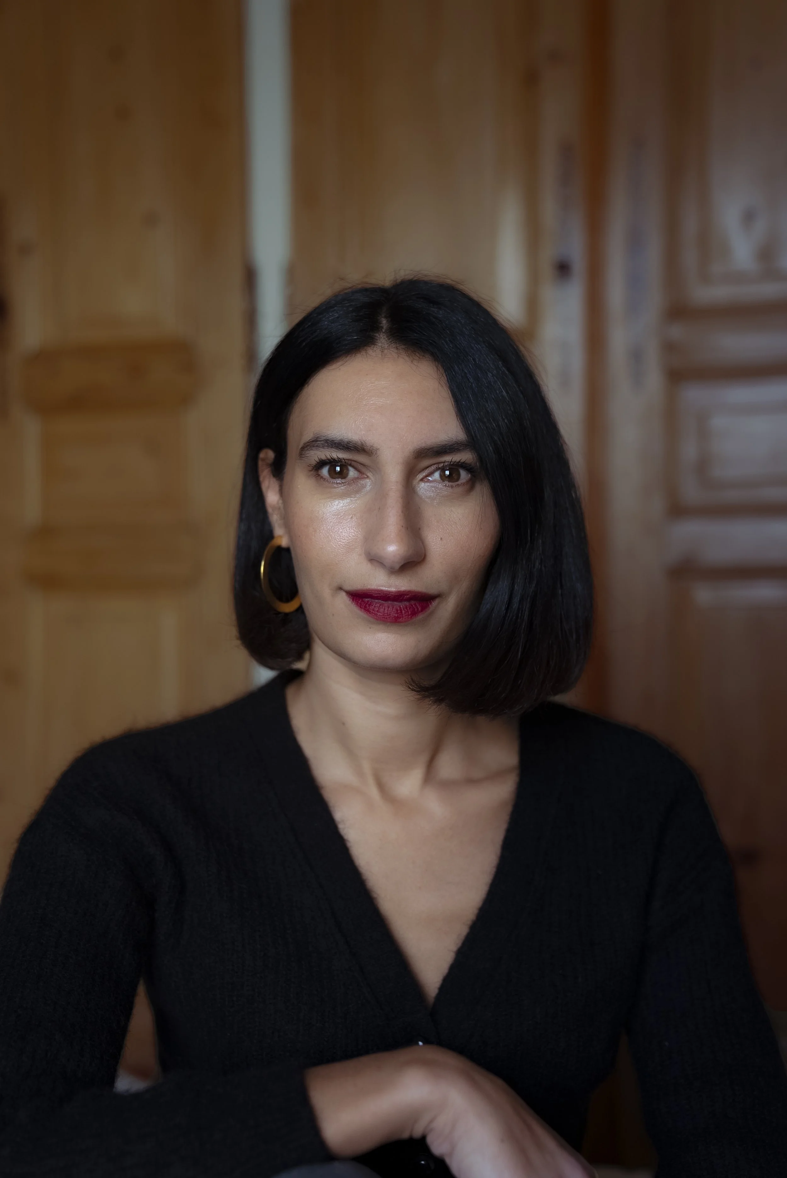 A woman with short black hair, wearing red lipstick, gold hoop earrings, and a black V-neck sweater, sitting against a wooden background.