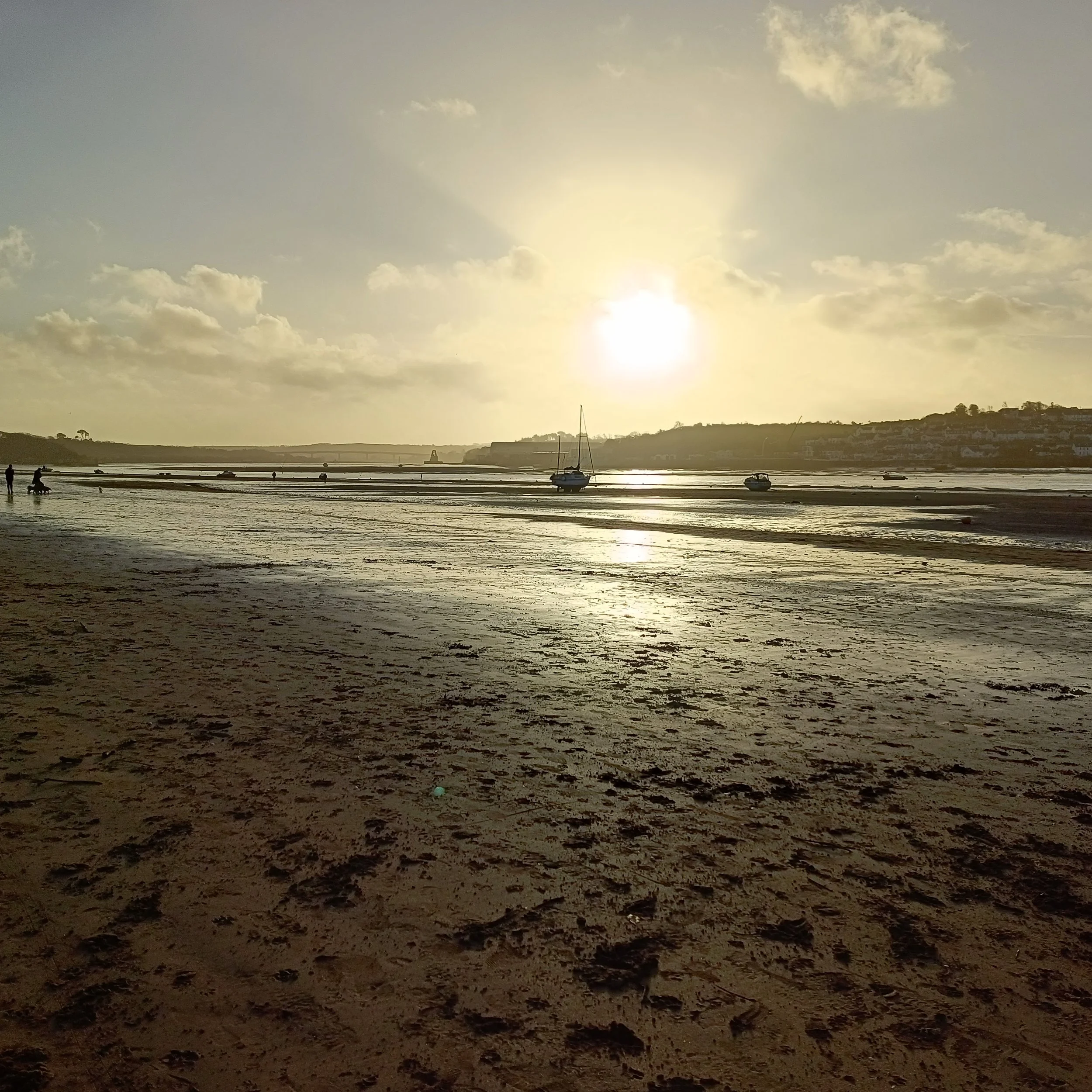 A sunset over a beach with boats anchored in the shallow water and people walking along the shoreline.