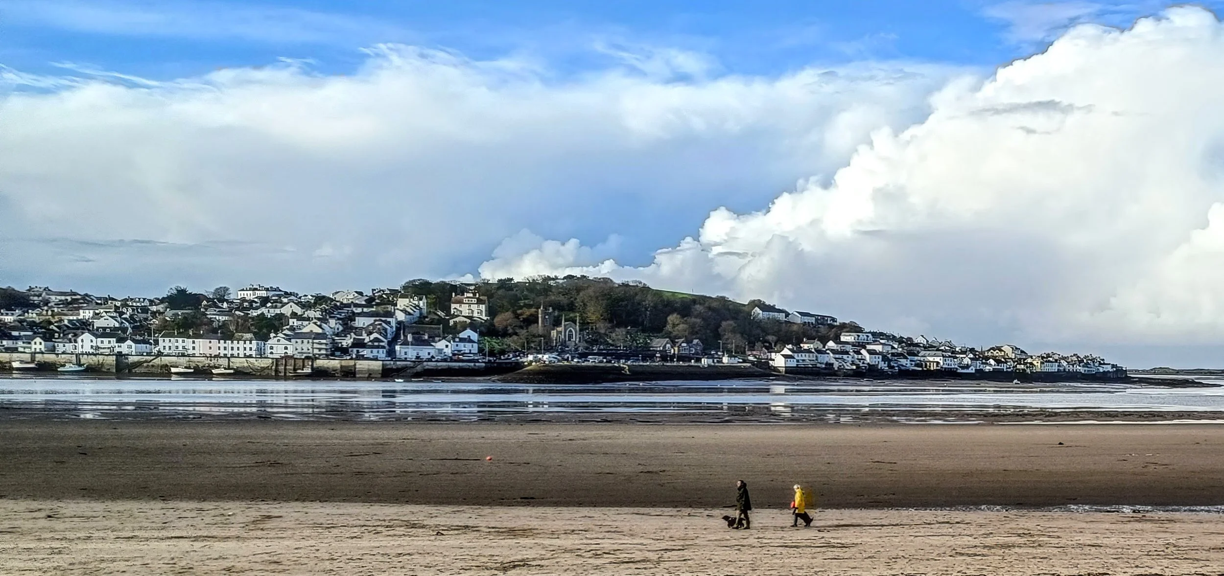 A coastal town with white houses on a hill, overlooking a sandy beach and calm water under a partly cloudy sky, with two people walking dogs along the shoreline.