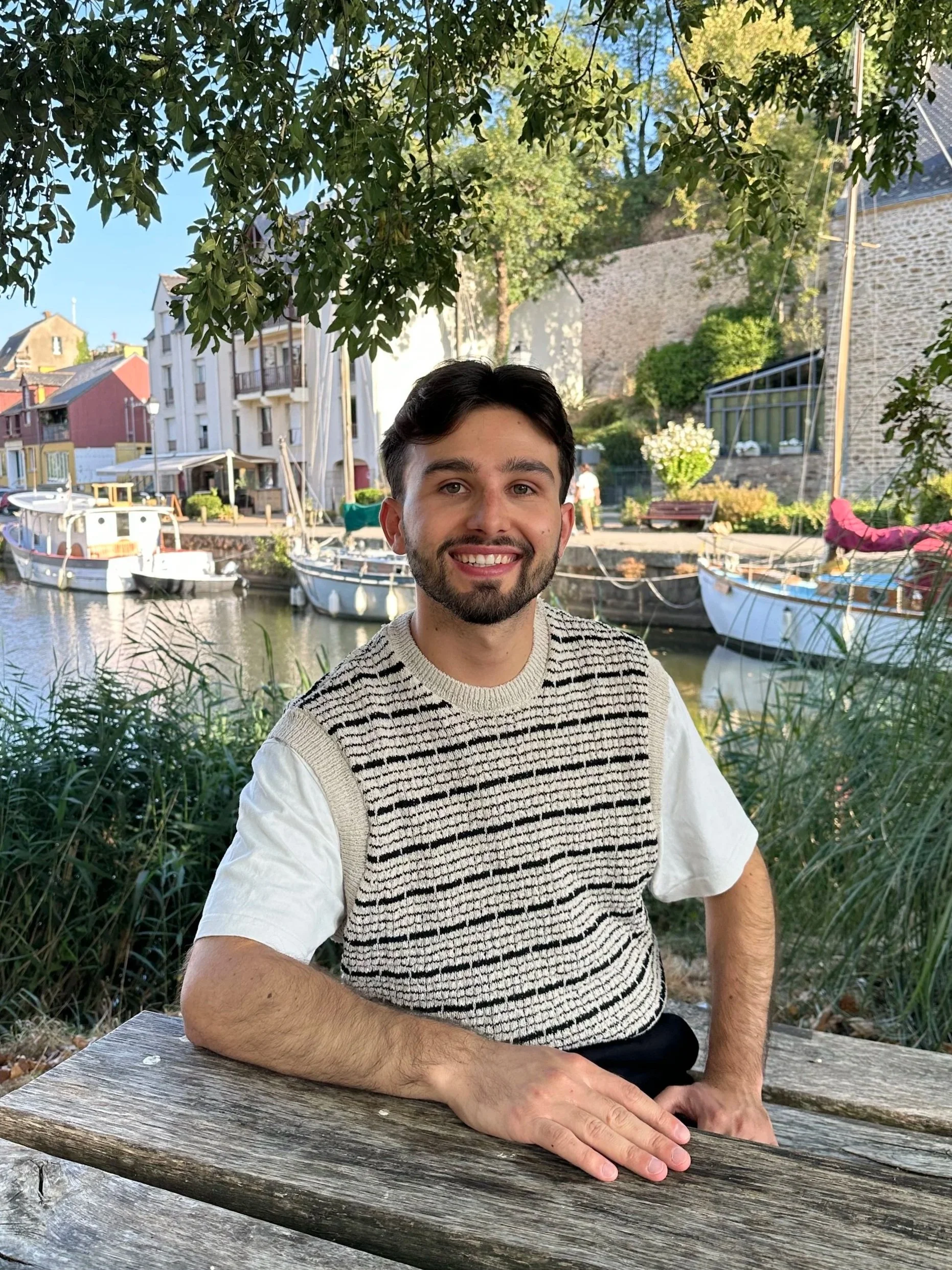 Un homme souriant assis à une table en bois à l'extérieur, avec des bateaux amarrés au port en arrière-plan et des maisons, sous un ciel bleu.