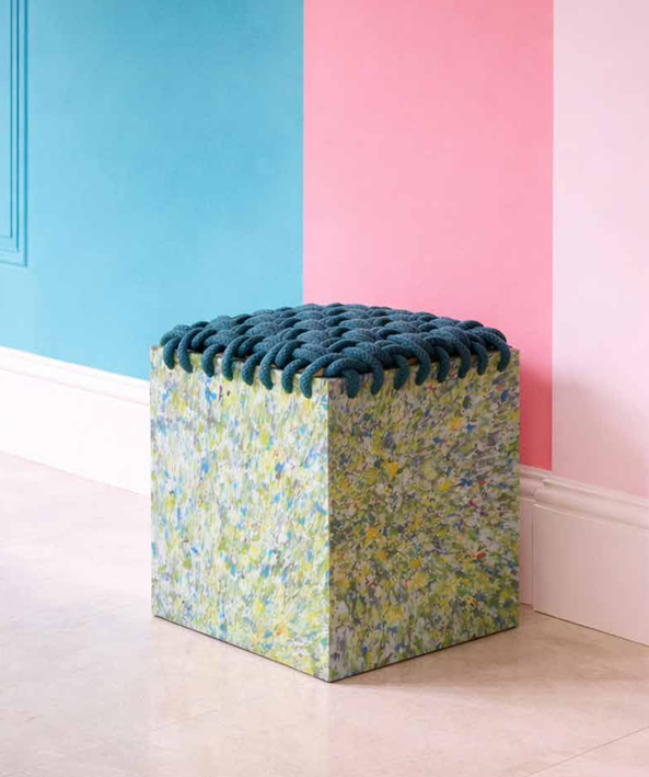 Un cube recouvert de peinture abstraite avec un coussin de tissu noué bleu sur le dessus, placé devant un mur bicolore bleu et rose.