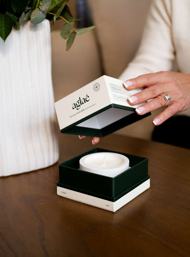 Une personne déballant une boîte contenant une bougie aromatique de la marque aglaé sur une table en bois, avec un vase blanc contenant des feuilles vertes à proximité.