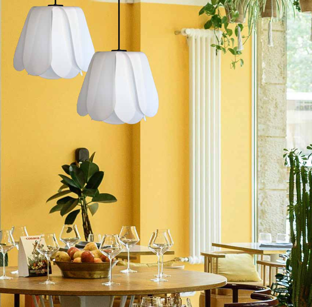 Salle à manger décorée avec deux suspensions blanches, un mur jaune, des plantes vertes, une table avec des verres à vin, une corbeille de fruits, et un fauteuil avec coussin, près d'une grande fenêtre.