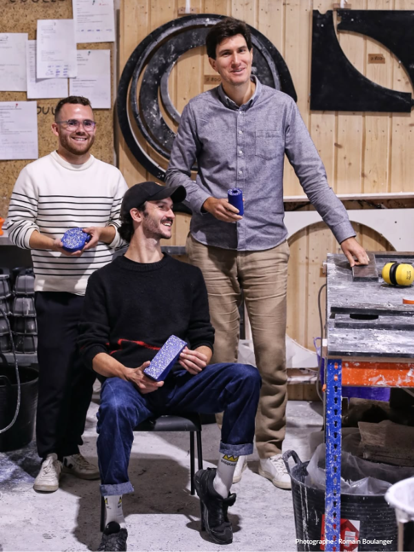 Trois hommes dans un atelier en train de célébrer, avec des cadeaux dans leurs mains, un fond en bois et des outils sur une table.